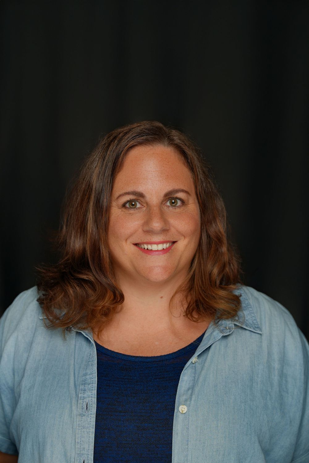 Professional headshot photo of someone smiling warmly at camera while wearing a light blue button-down shirt over navy top.