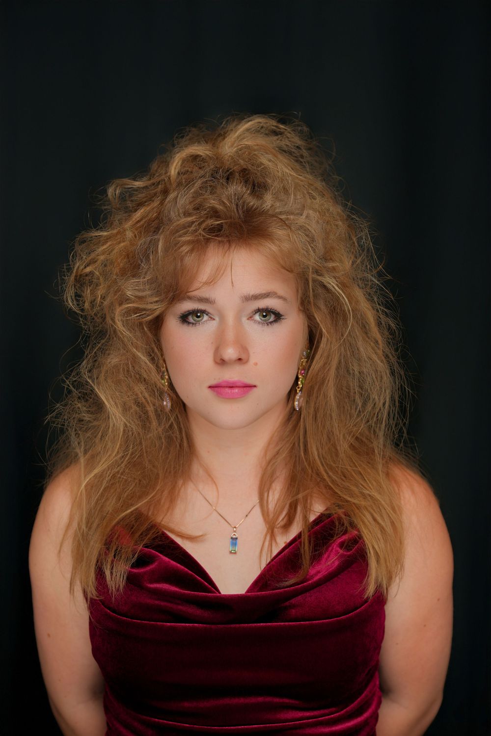 A portrait of someone in a burgundy velvet dress with voluminous honey-blonde hair against a dark background.