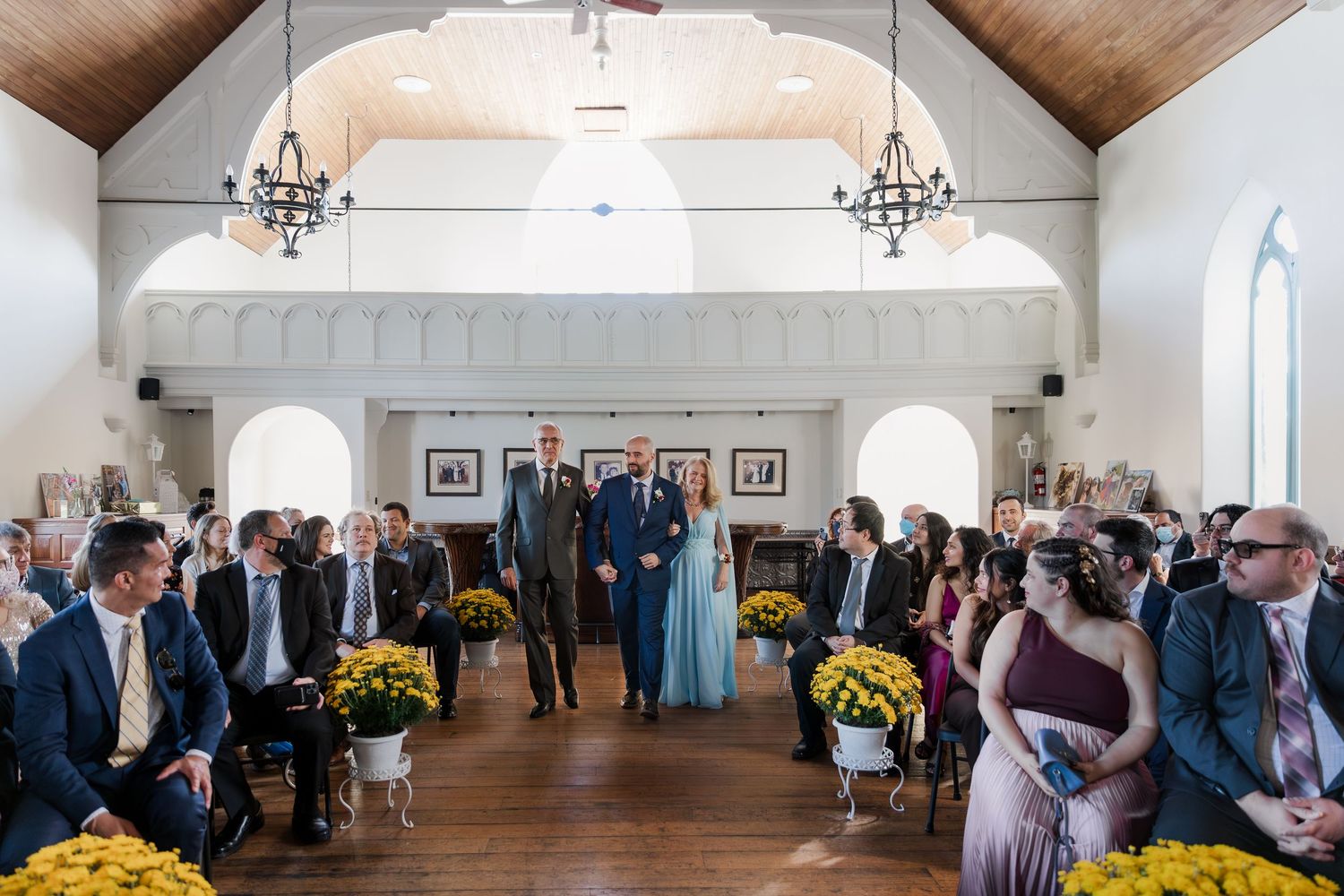 Groom walks down the aisle with his parents in the chapel room surrounded by guests and yellow florals at Hauser Hall in Kitchener, Ontario.
