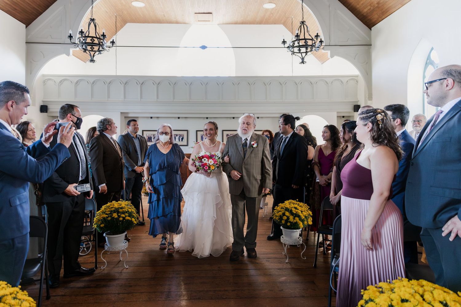 Bride walks down aisle with parents in rustic chapel with vaulted ceilings, chandeliers and yellow florals at Hauser Hall in Kitchener.