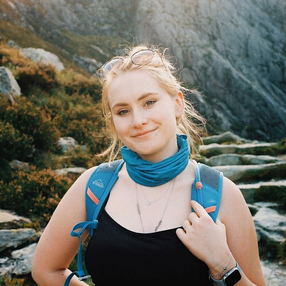A hiker wearing a blue neck gaiter and backpack smiles while on a rocky mountain trail in warm sunlight.