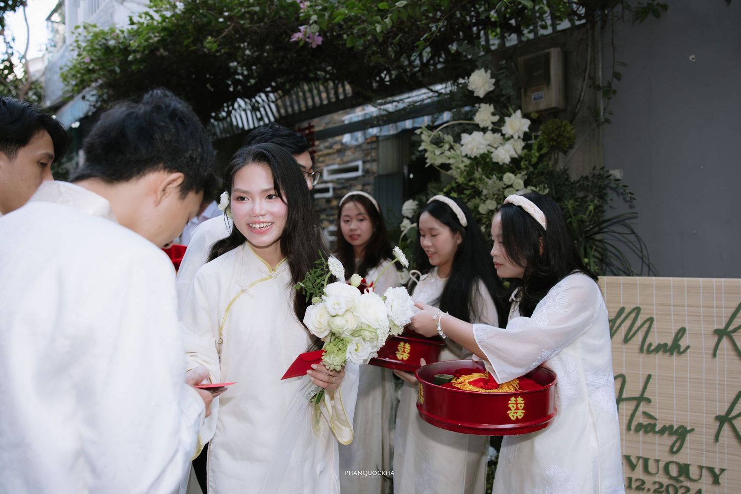Smiling bride in white ao dai receives bouquet during Vietnamese traditional wedding ceremony.