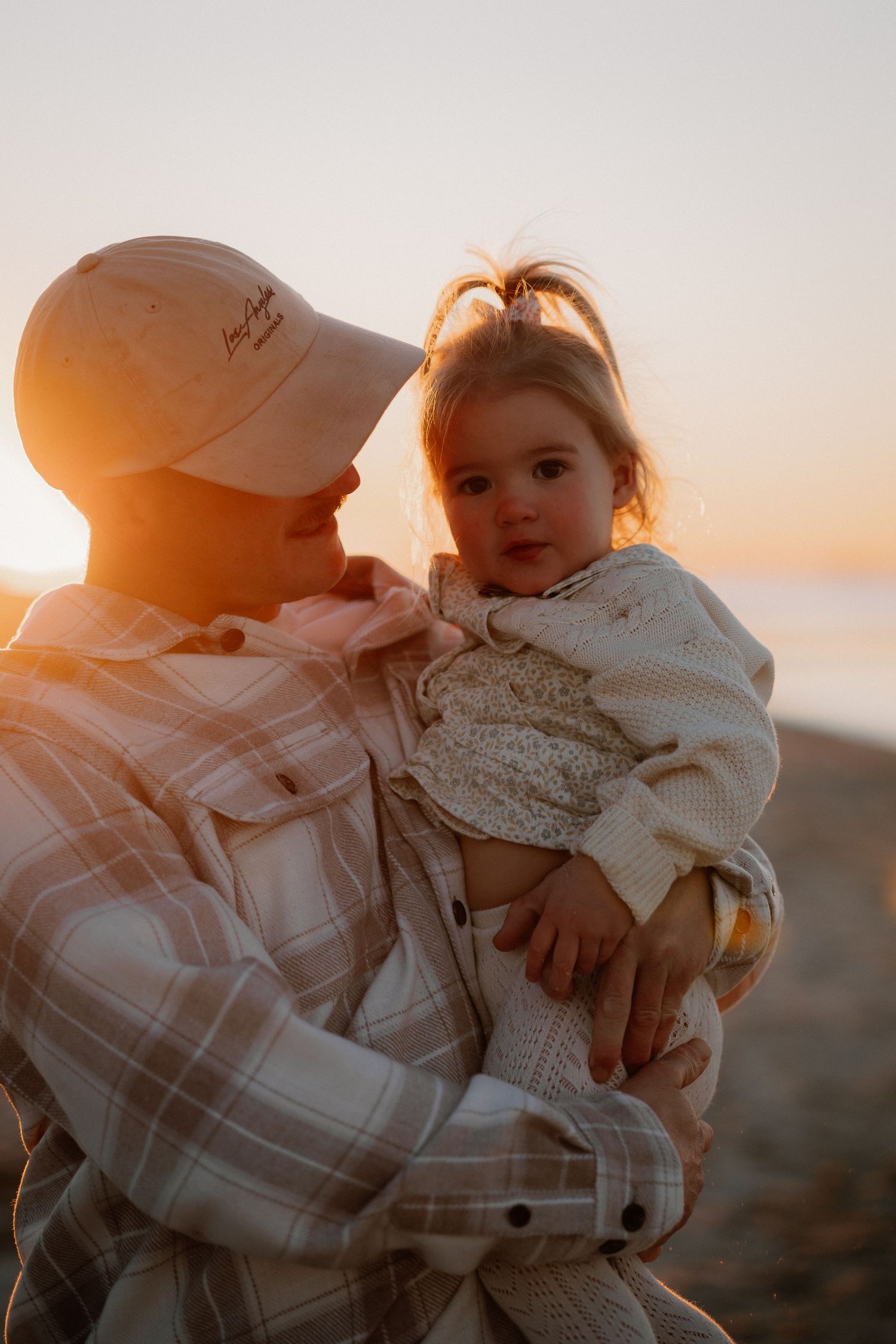 A tender sunset moment between parent and child in a soft knit sweater and plaid shirt against a glowing warm sky.
