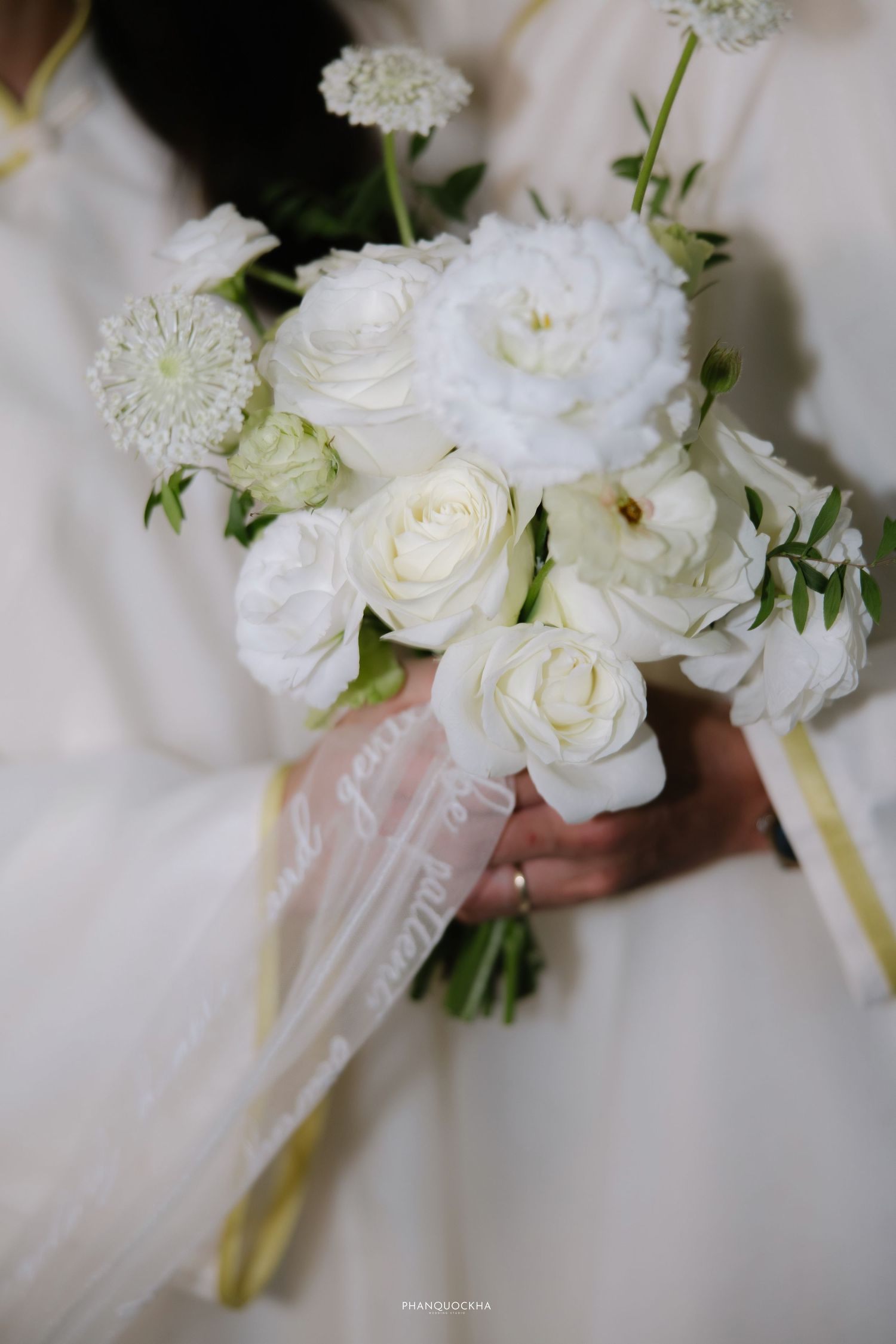 Elegant bridal bouquet of white roses, ranunculus, and scabiosa with sheer ribbon held by bride in white ao dai.