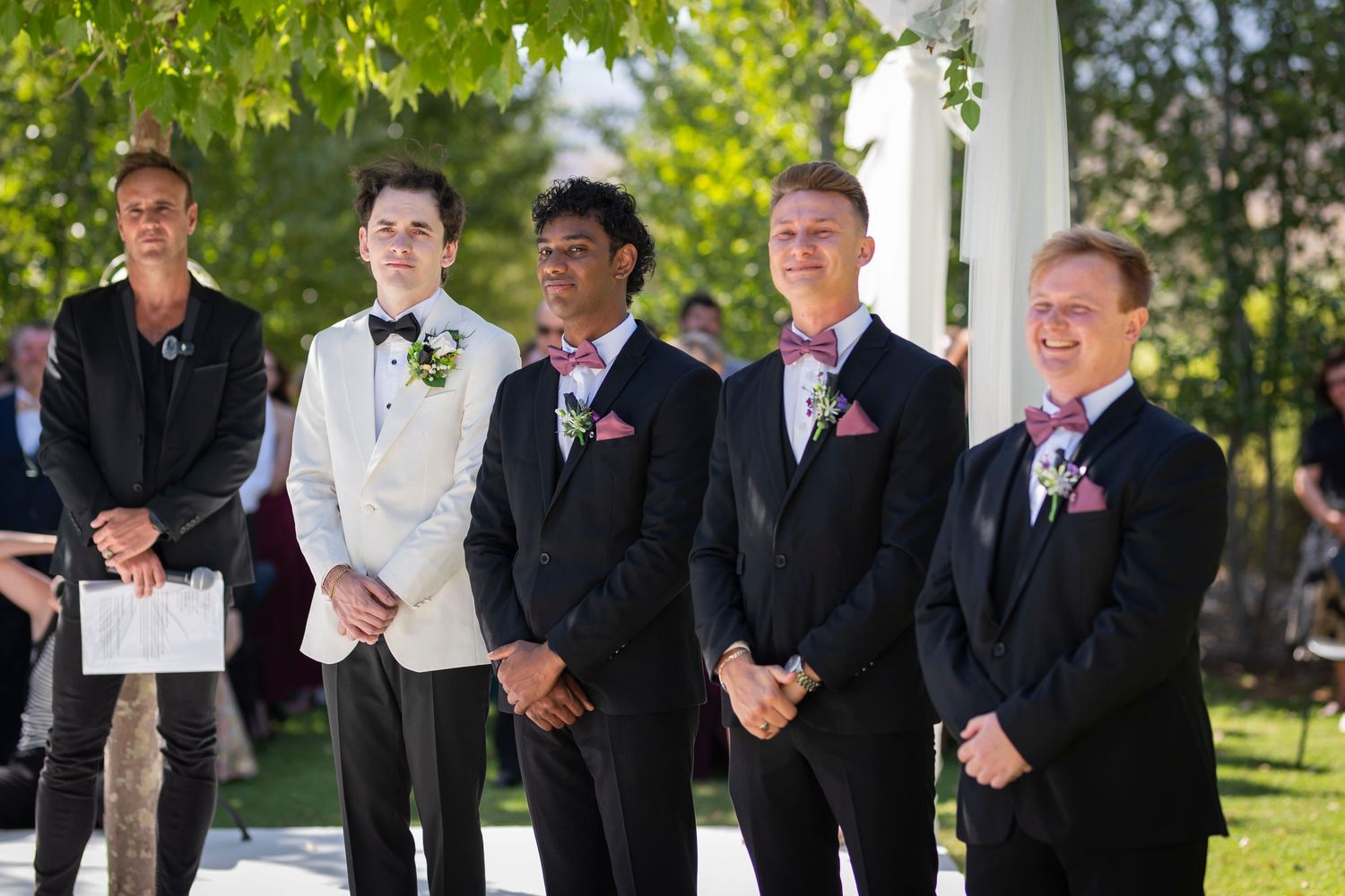A group of groomsmen stand together under a white wedding arch surrounded by green leaves during an outdoor ceremony.