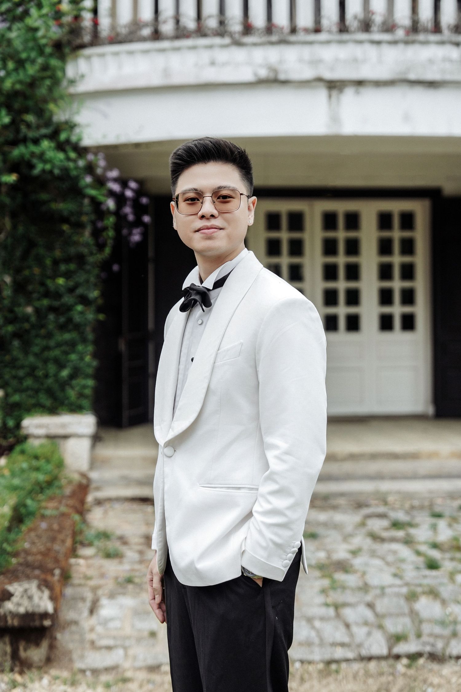 Stylish Asian groom in white tuxedo jacket with black bow tie and glasses posing outdoors at wedding venue.