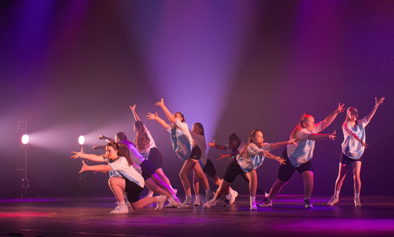 Dancers perform an energetic contemporary dance routine on stage with dramatic purple lighting and dynamic poses.