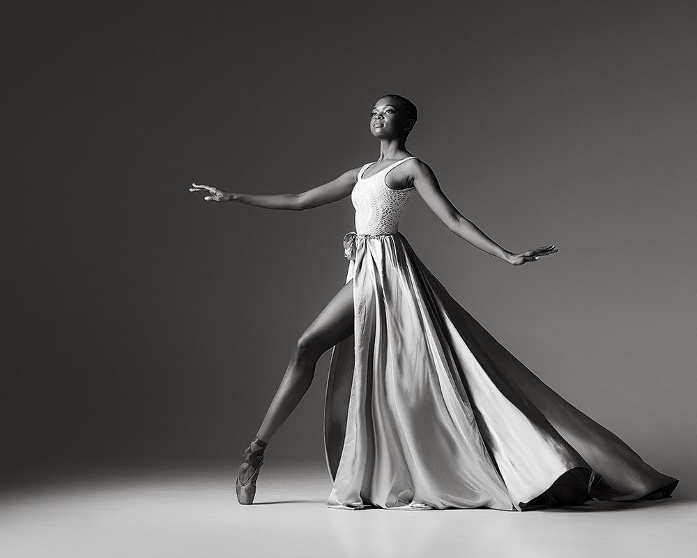 A ballet dancer in a light flowing dress strikes an elegant pose with extended arms against a dark background.