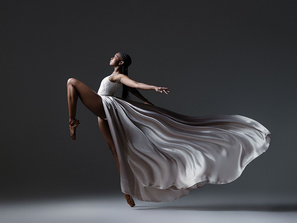 A figure in flowing white fabric performs a graceful dance movement across a dark studio setting.