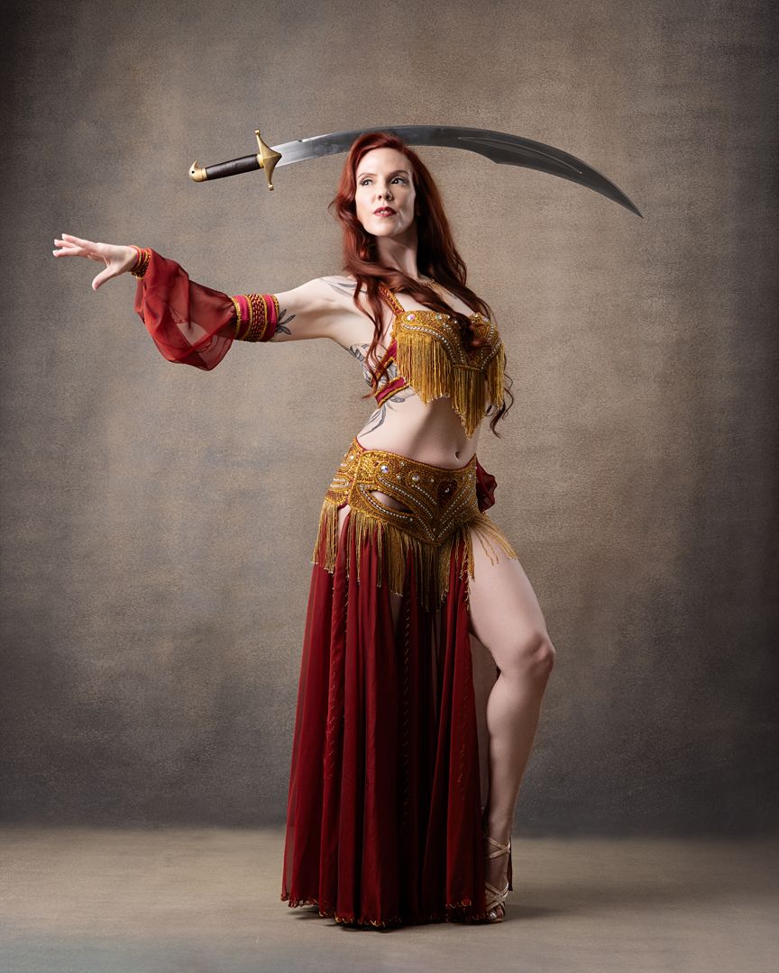 Red-haired woman in gold and red belly dance costume wielding a large curved sword in a dramatic pose.
