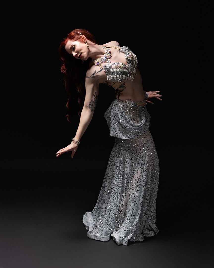 Red-haired woman in a sparkling silver belly dance costume poses gracefully against a dark background.