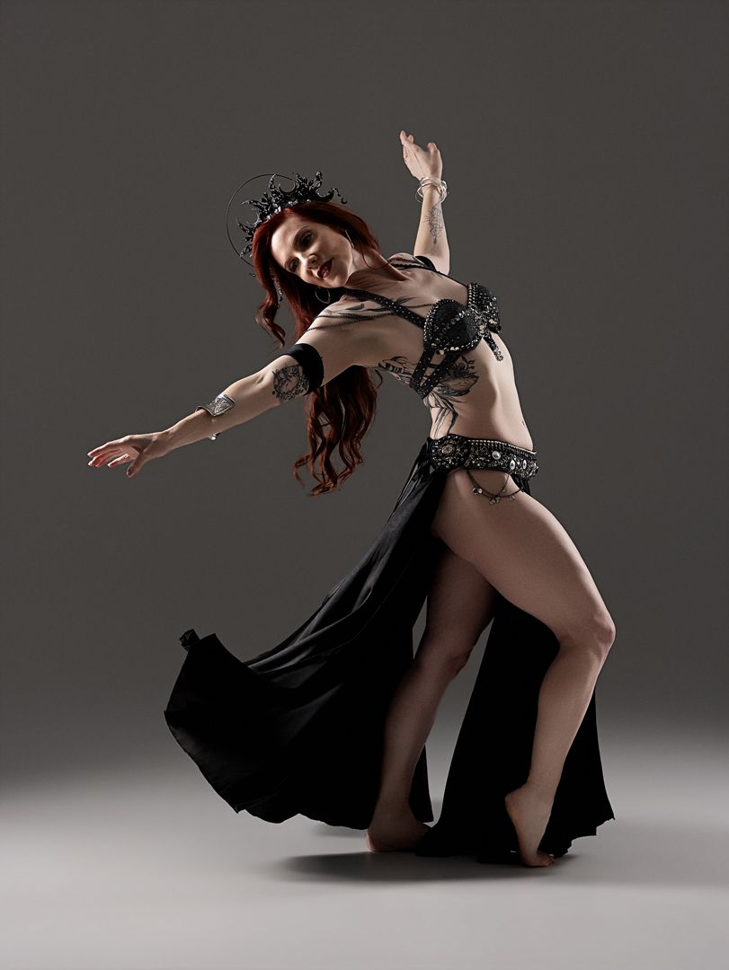 A belly dancer in a black costume performs an expressive pose against a gray studio background.
