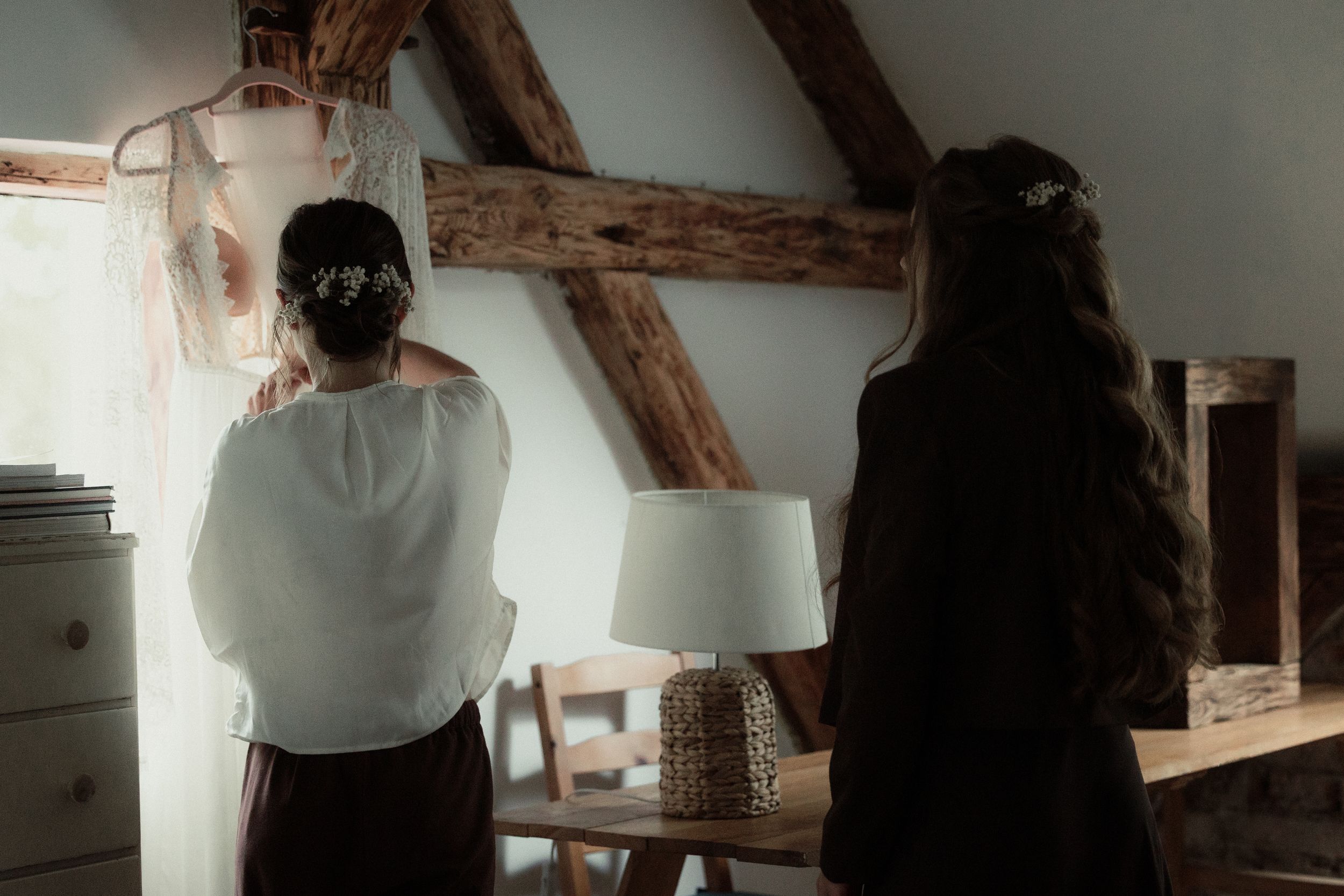 Two people stand in rustic room with exposed wooden beams and furniture, photographed from behind in dim lighting.