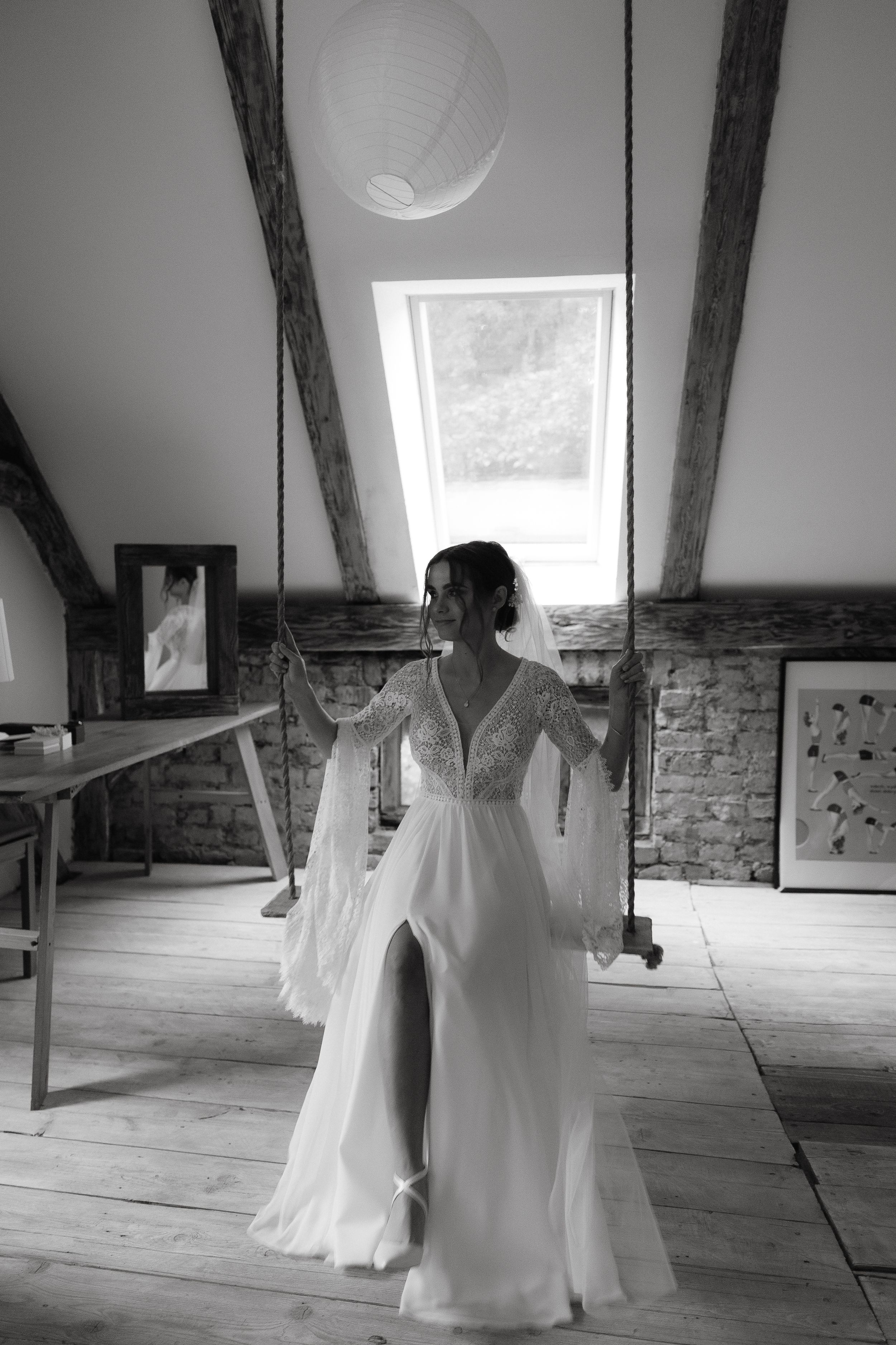 Black and white photo of a wedding dress with flowing fabric and lace details in a rustic attic room with exposed beams.