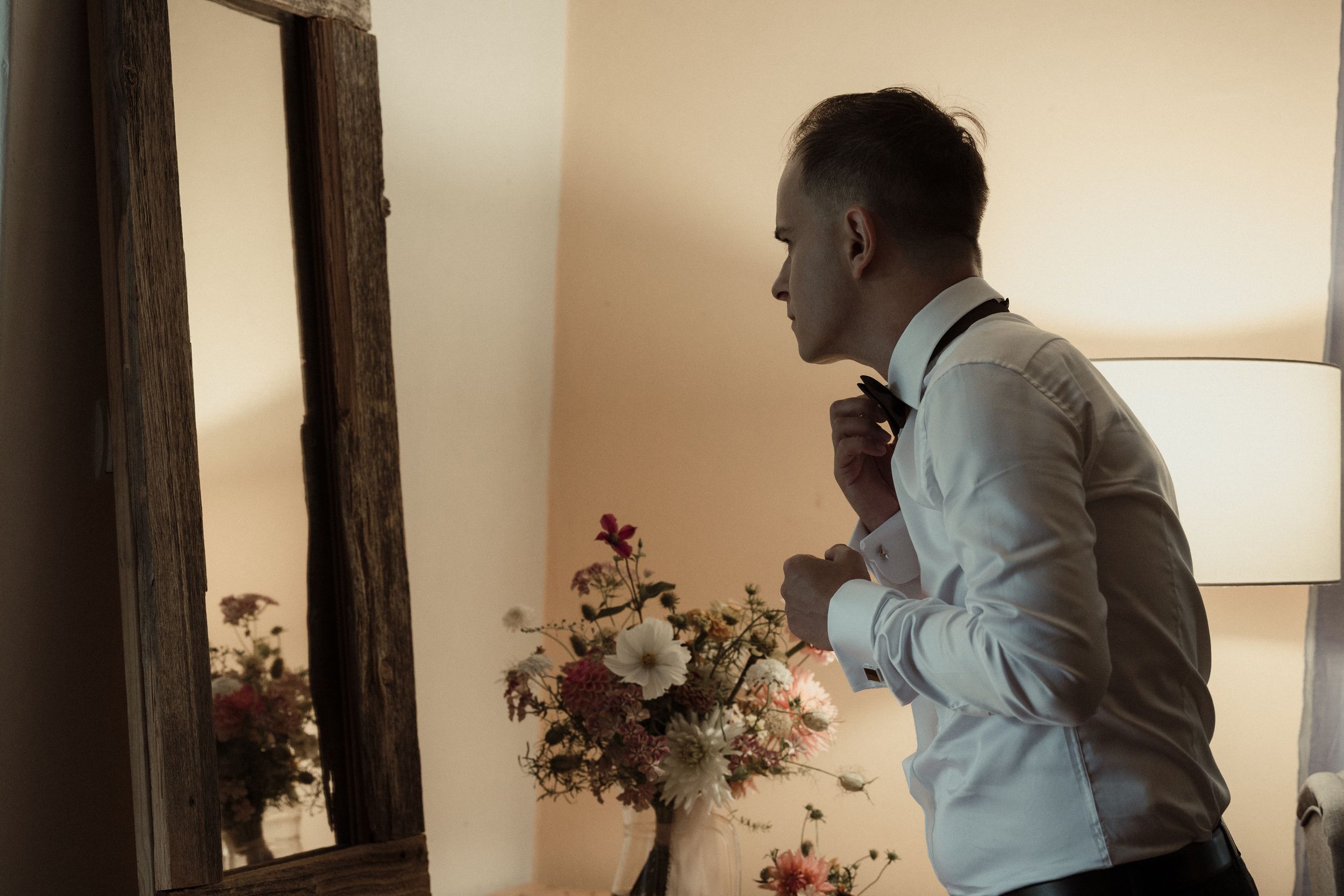 Someone in a white dress shirt and bow tie adjusts their attire while standing near a mirror and floral arrangement.