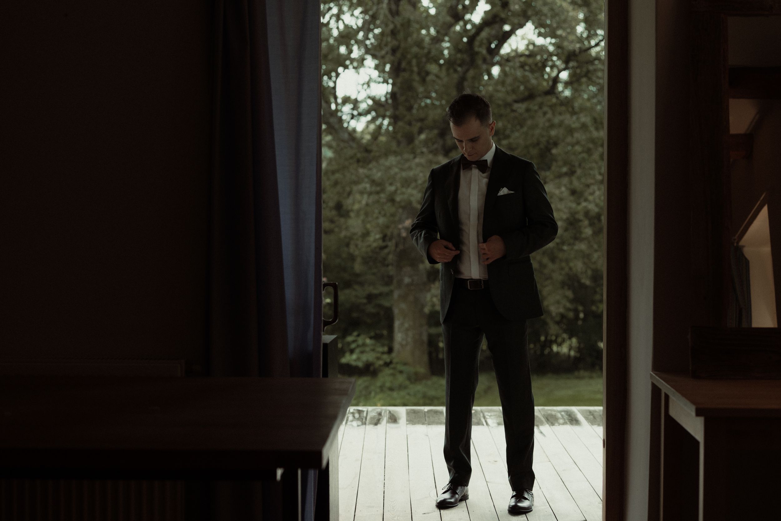 A person in a dark formal suit stands in a silhouette against bright daylight on a wooden deck or porch.