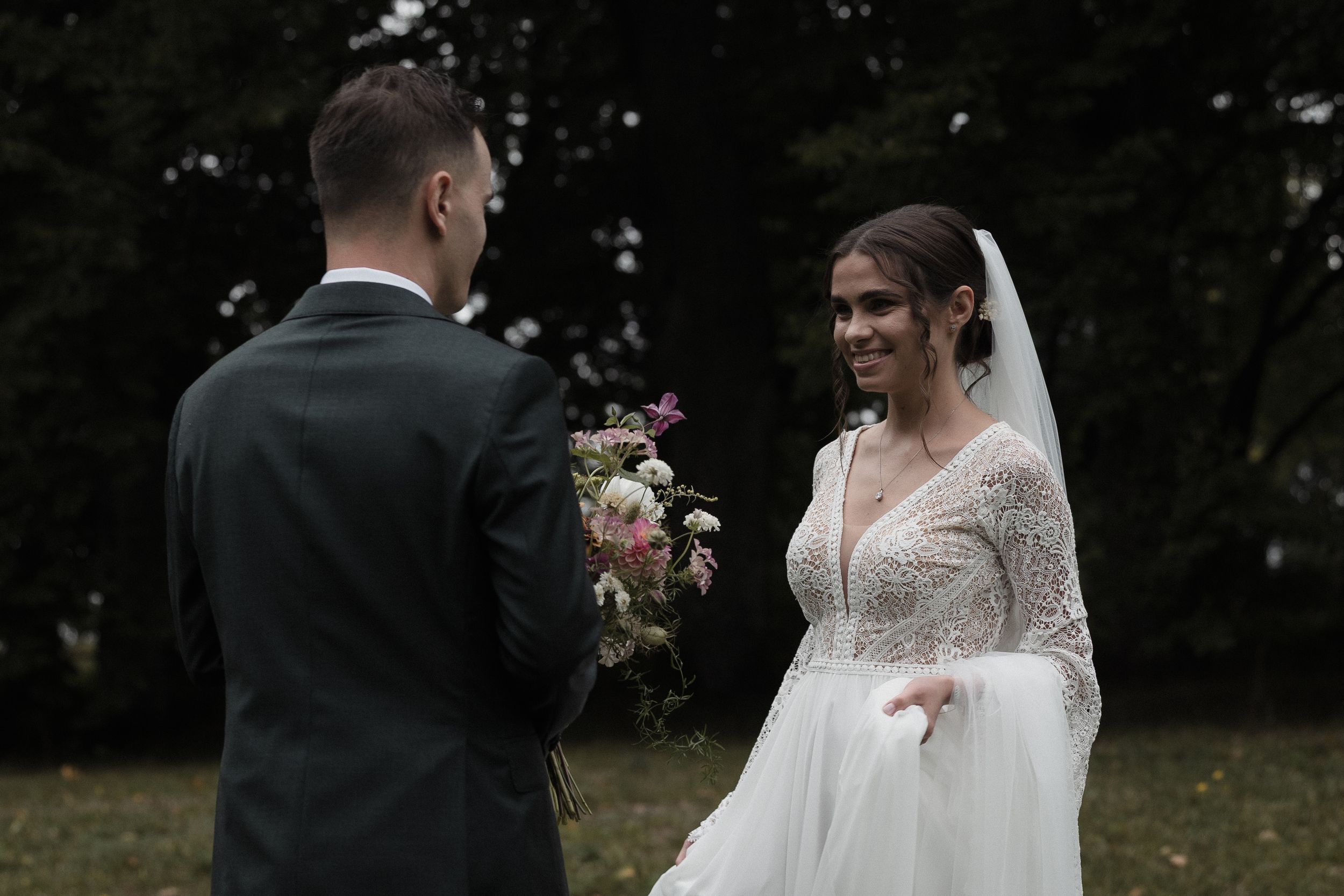 A romantic outdoor wedding ceremony in a lush park setting with the couple exchanging rings under a canopy of trees.