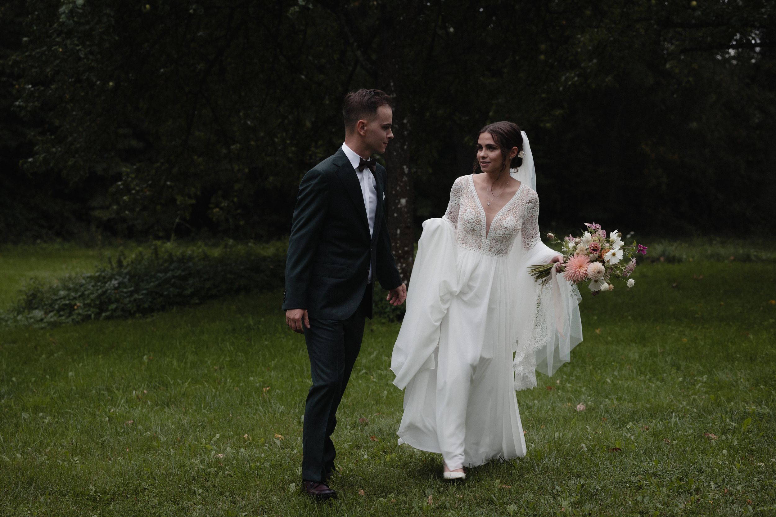 A romantic wedding couple walks hand in hand through a lush green garden path at an outdoor ceremony.