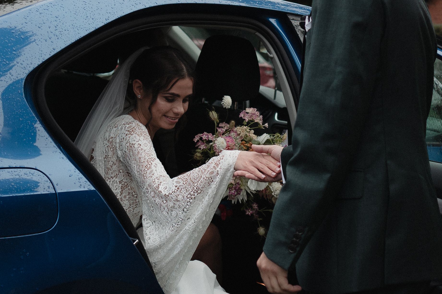 A bride and groom exit their wedding ceremony in classic vintage car as guests throw flower petals in celebration.