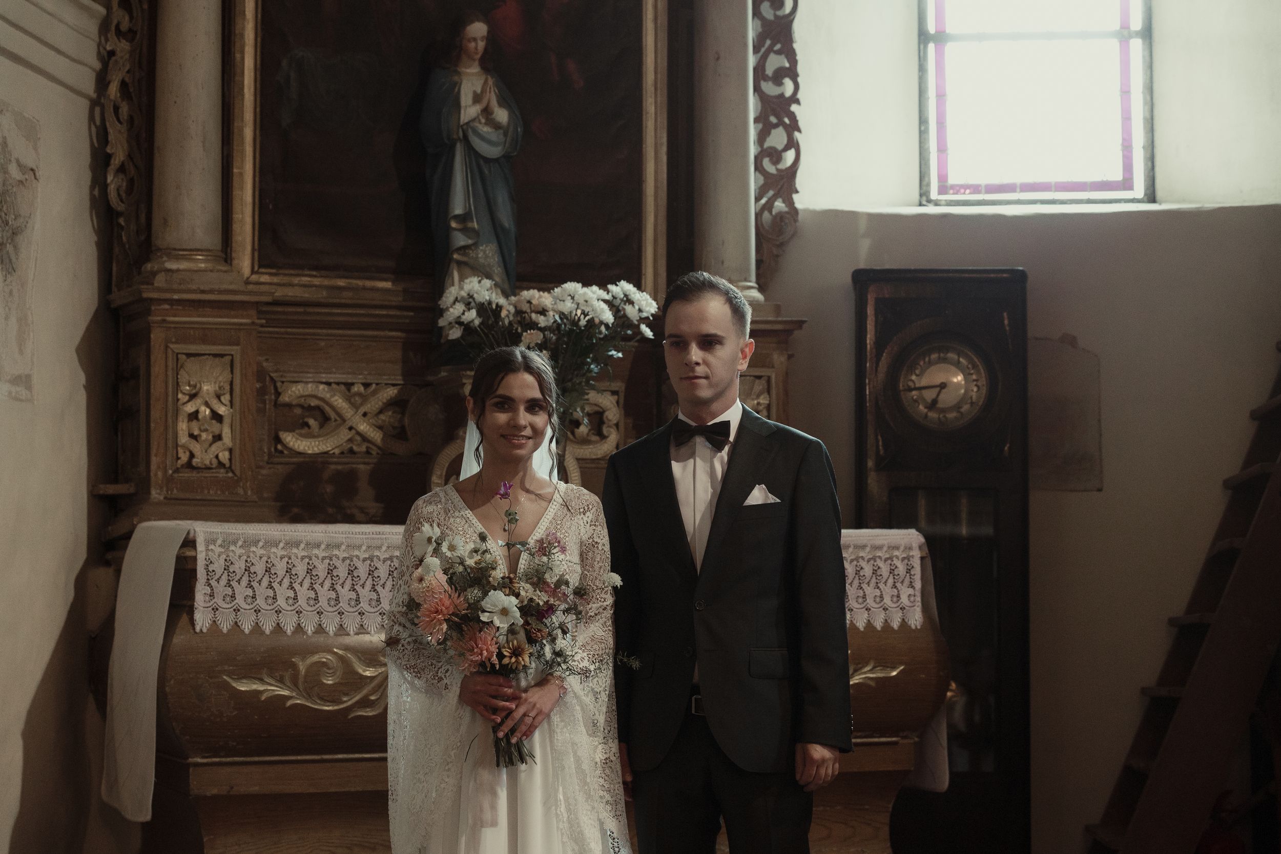 Elegant wedding couple stands together in a traditional church with ornate decor and soft natural lighting.
