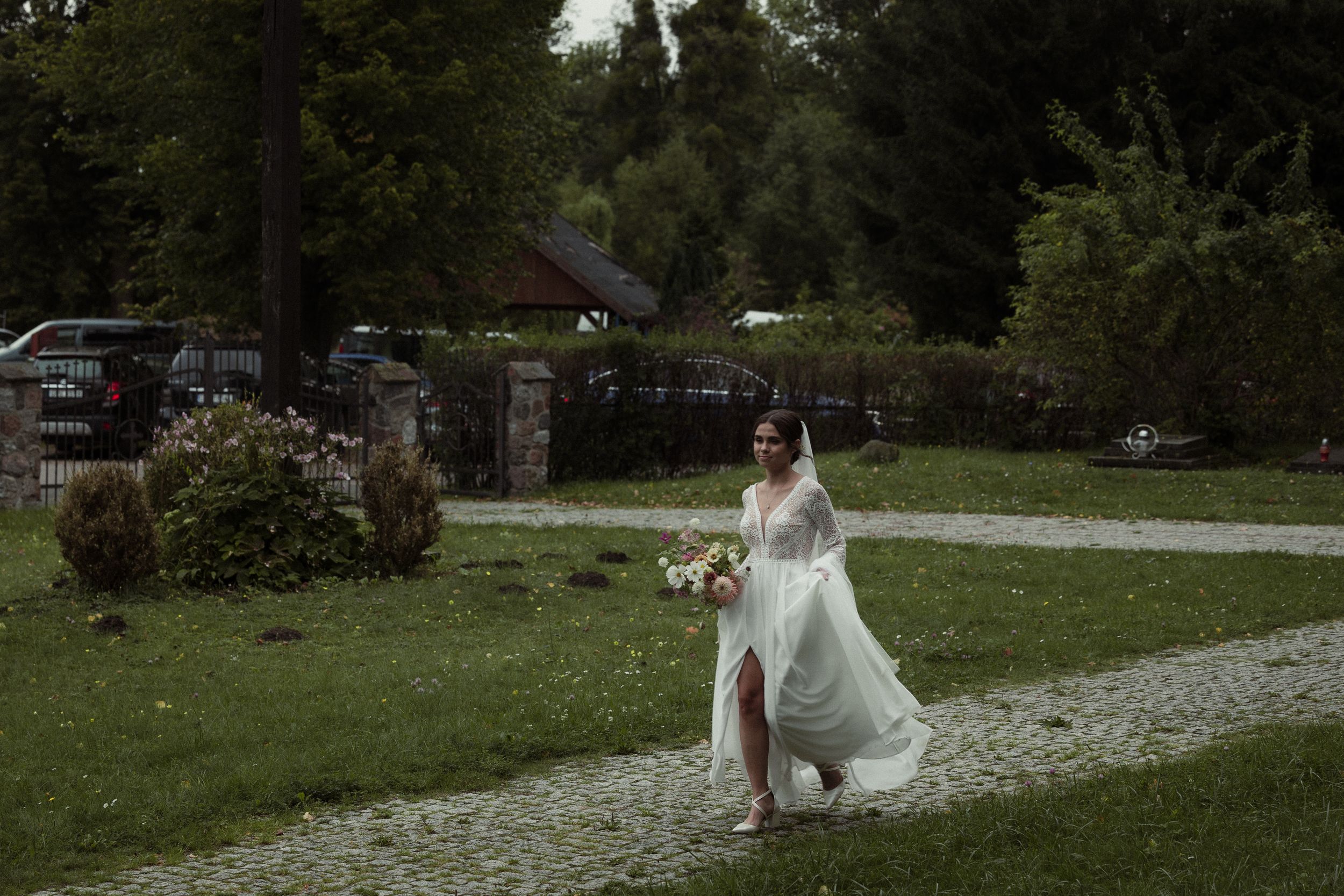 A bride in a flowing white dress and long veil walks down a garden path surrounded by lush greenery.