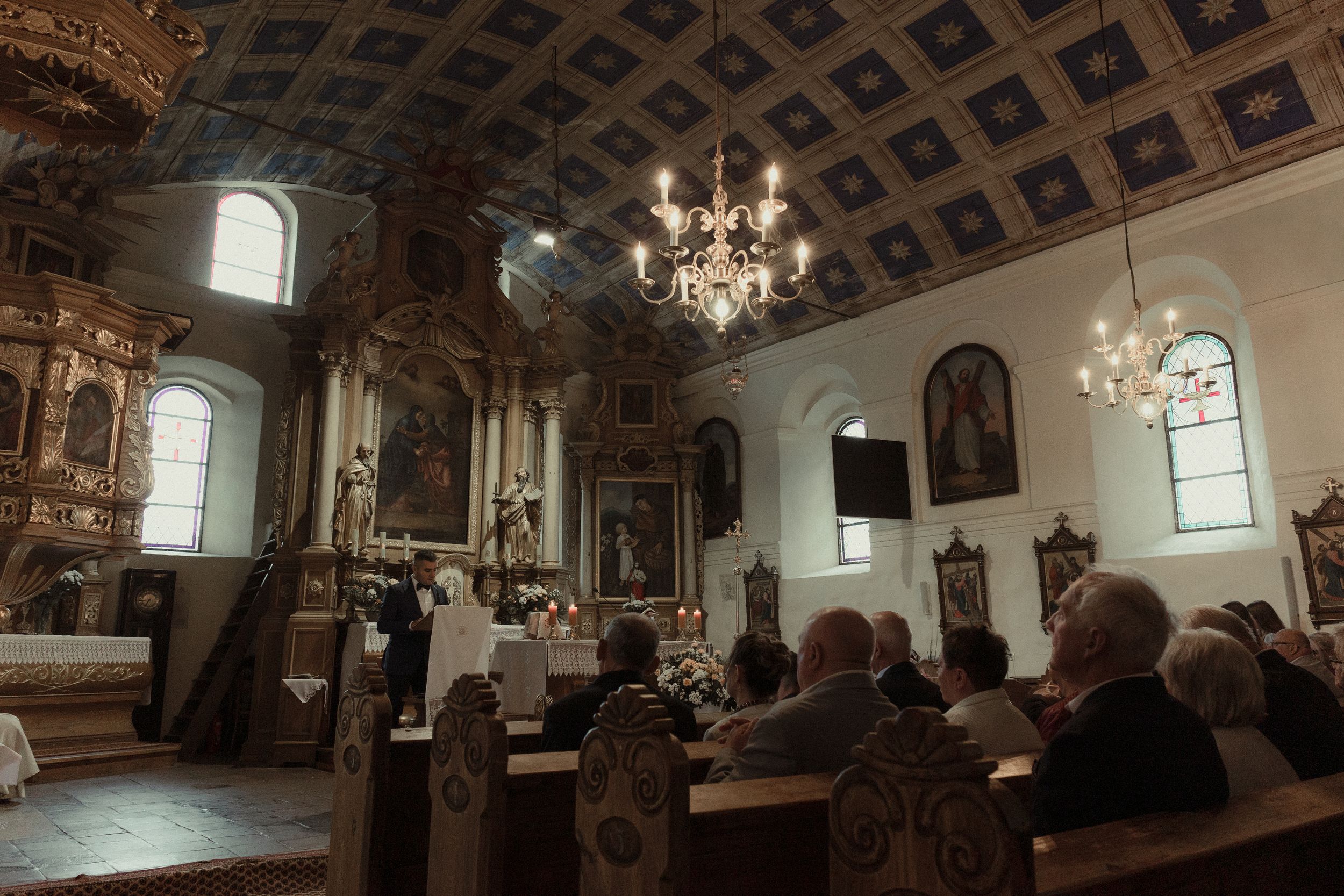 Ornate church interior with coffered ceiling, wooden pews, and hanging chandeliers bathed in natural light.