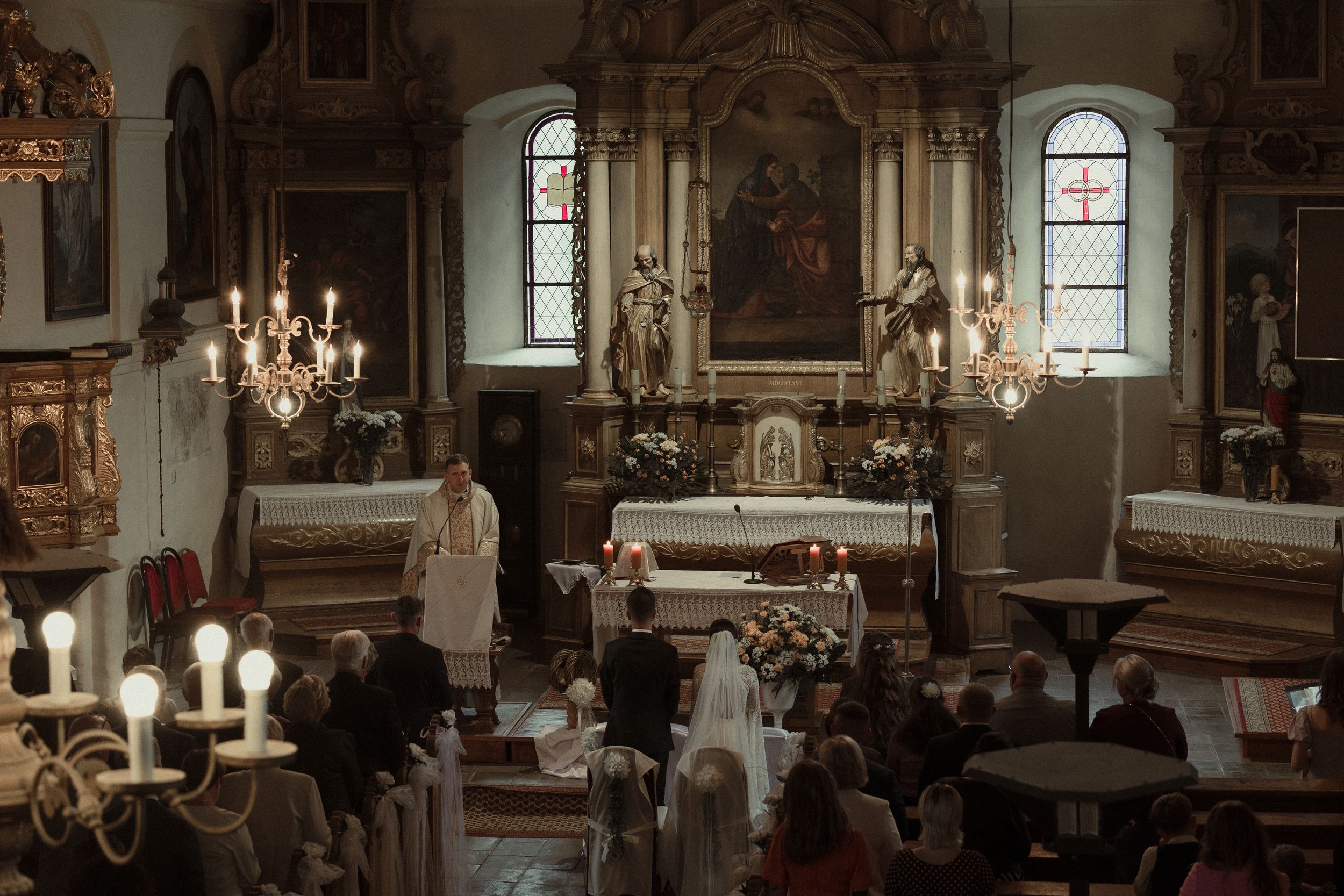 Traditional wedding ceremony taking place in an ornate church sanctuary with candelabras and stained glass windows.