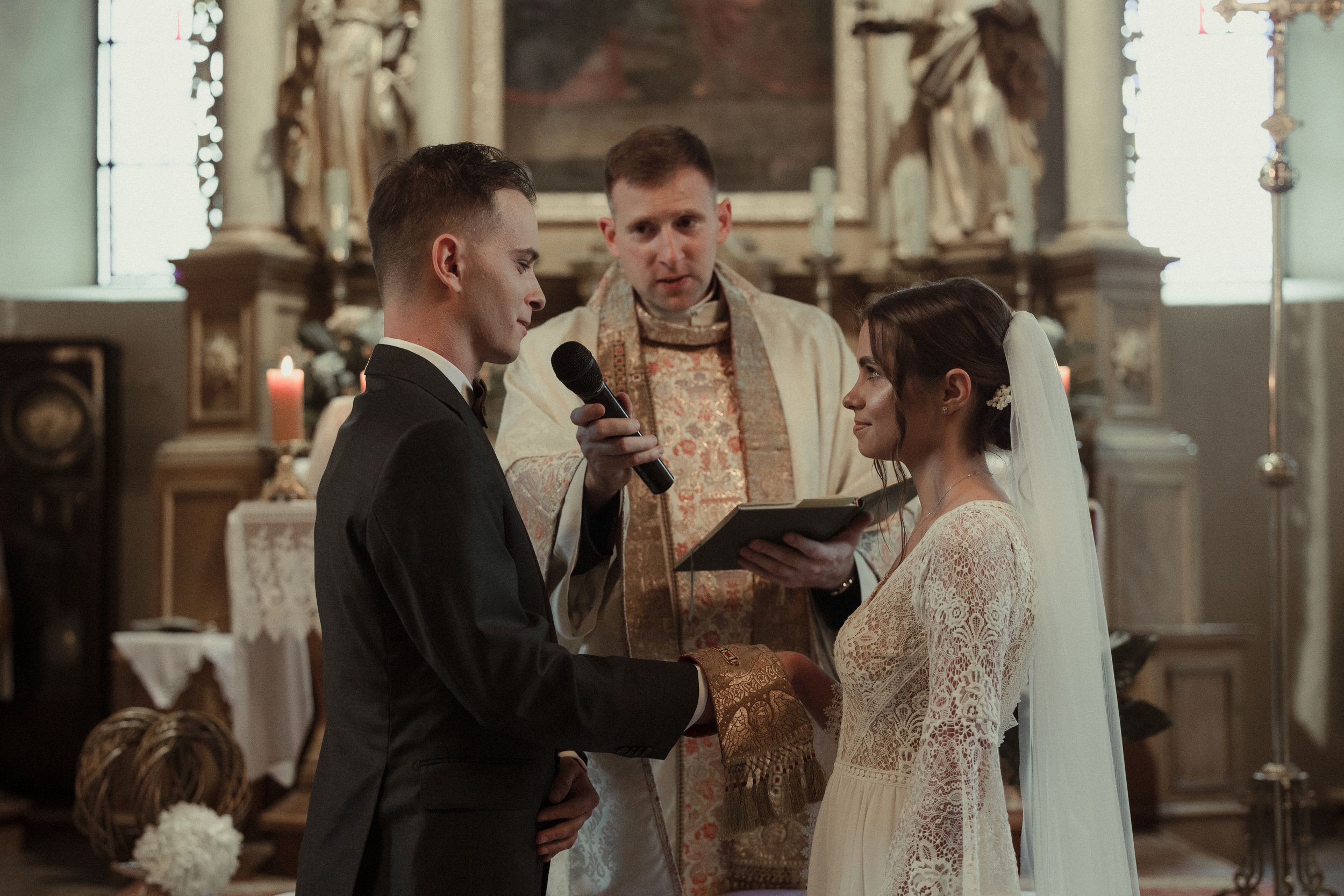 A couple in formal wedding attire exchanges rings during their ceremony inside a sunlit church.