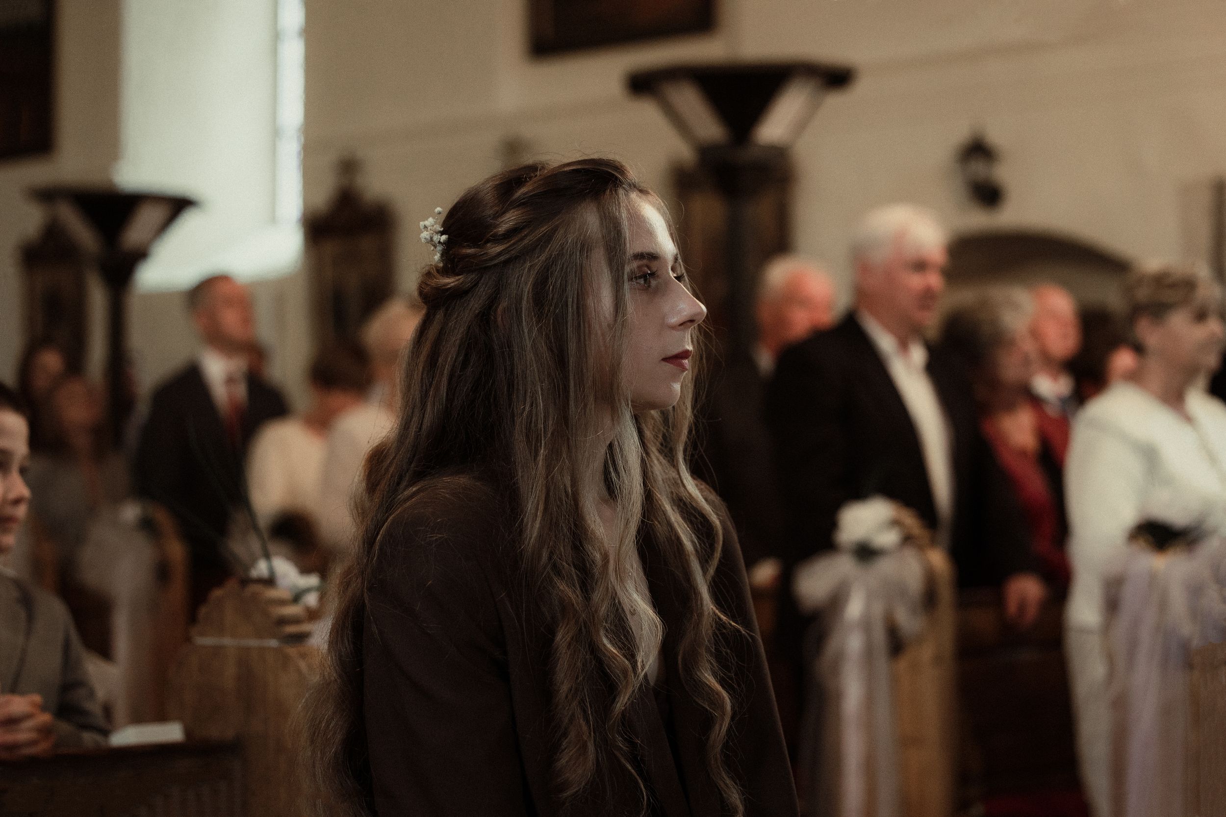 A person with long wavy brown hair stands in a dimly lit church gathering with other attendees in the background.