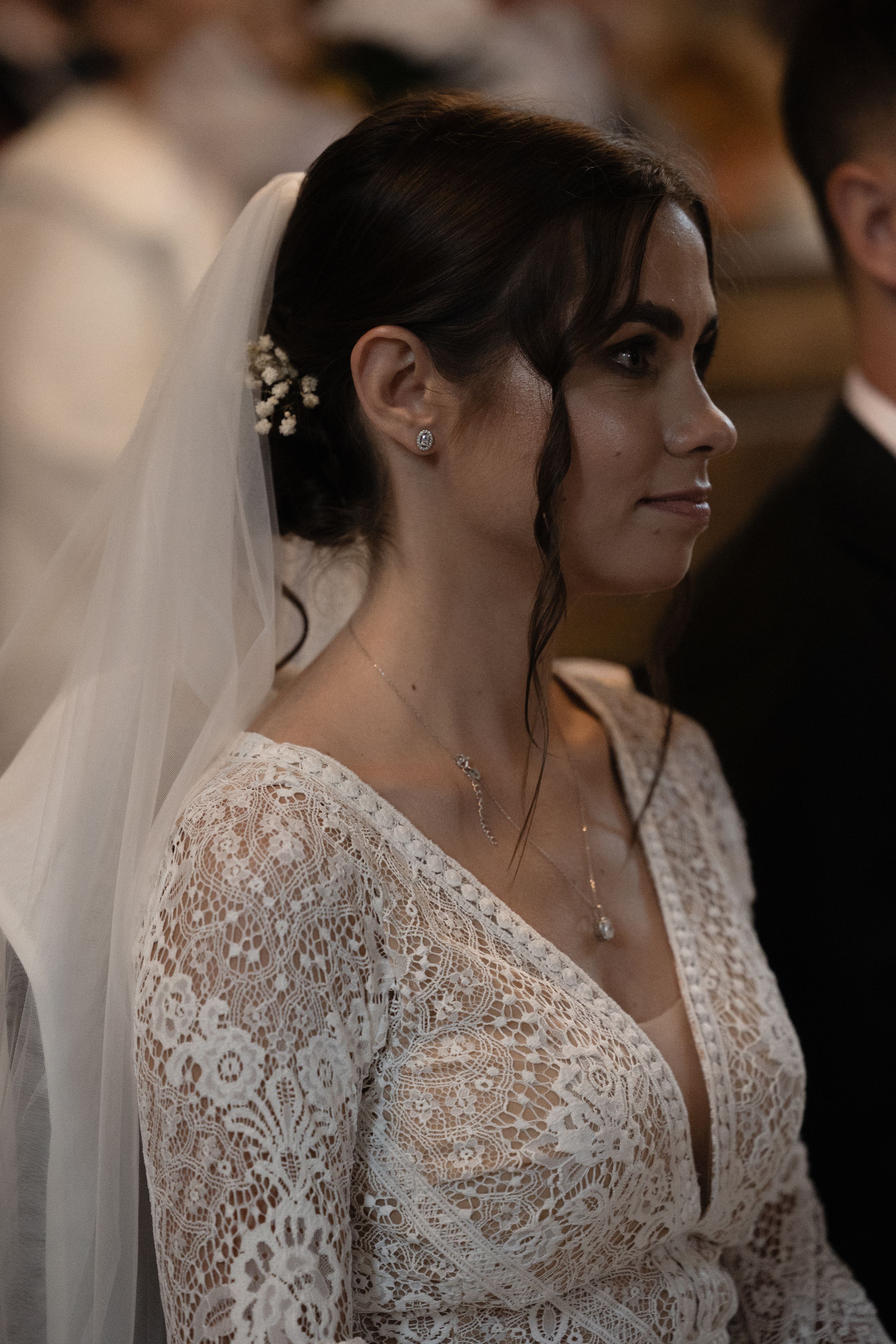 A bride wearing a delicate lace white wedding dress and veil is shown in profile during her special day.