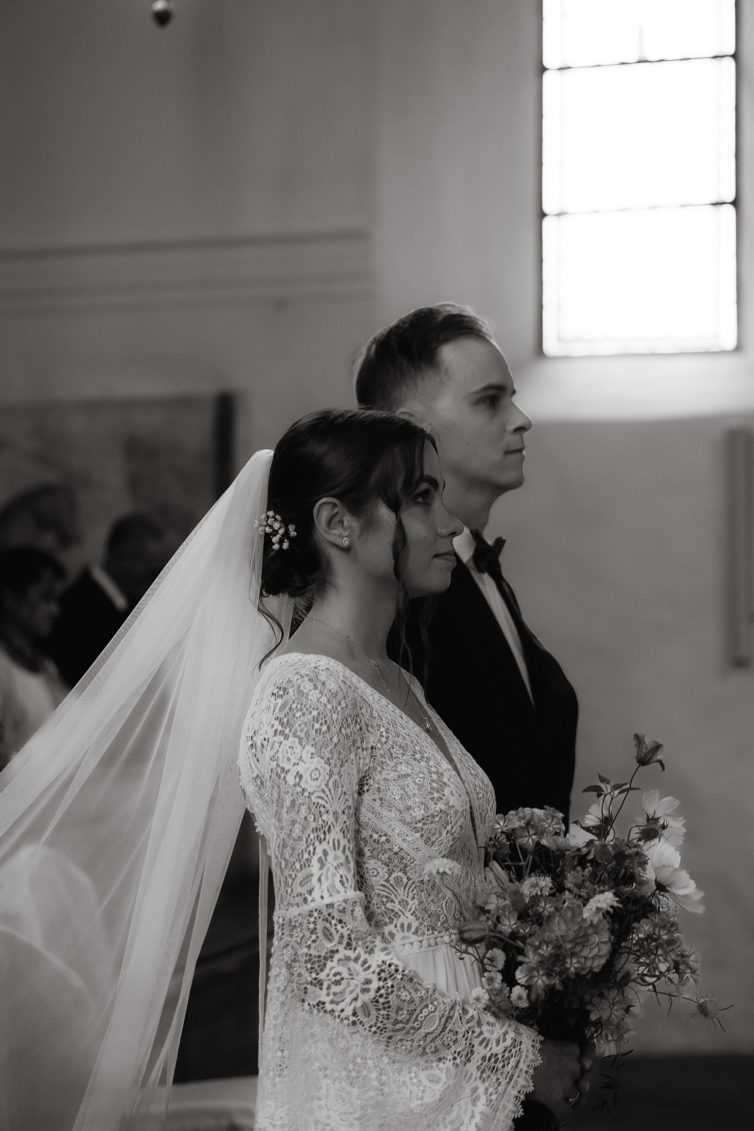 A bride and groom stand side by side in a church during their wedding ceremony with a flowing white veil.