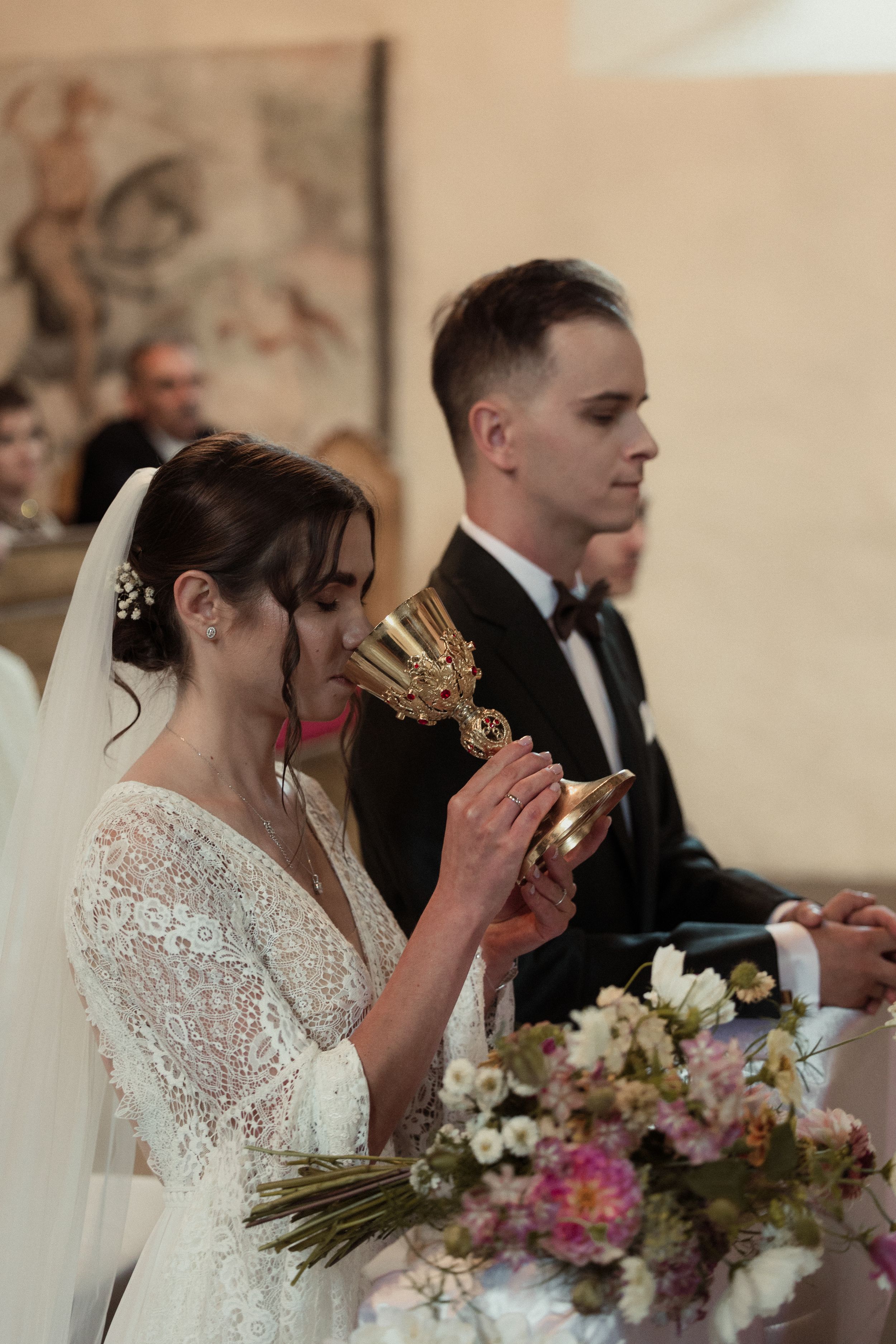 Bride in white lace dress drinking from chalices during wedding ceremony among floral arrangements.