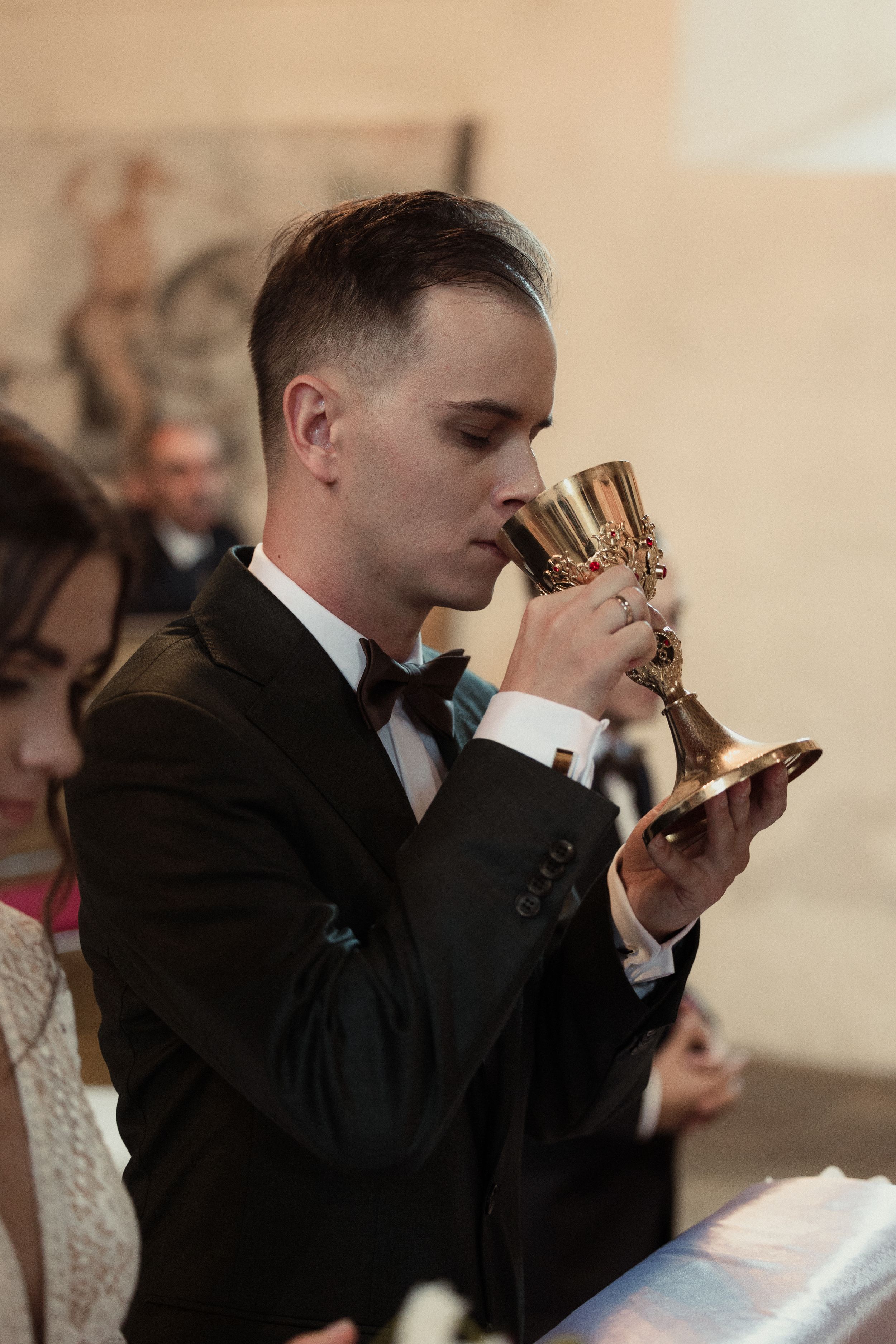 Groom in black suit drinking from communion cup during traditional wedding ceremony.