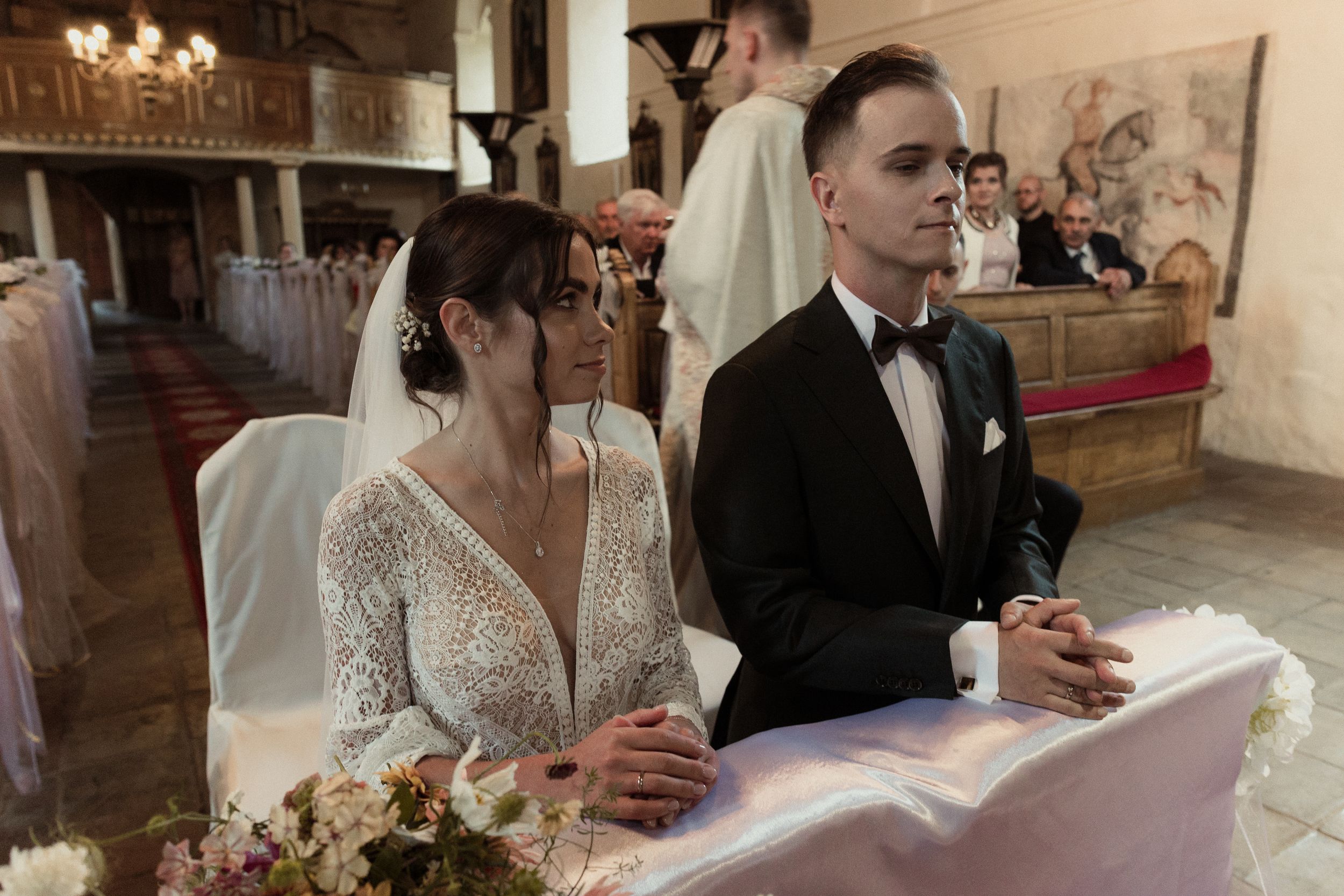 A couple sits at a reception table while signing papers, with the bride wearing a lace wedding dress in an elegant venue.