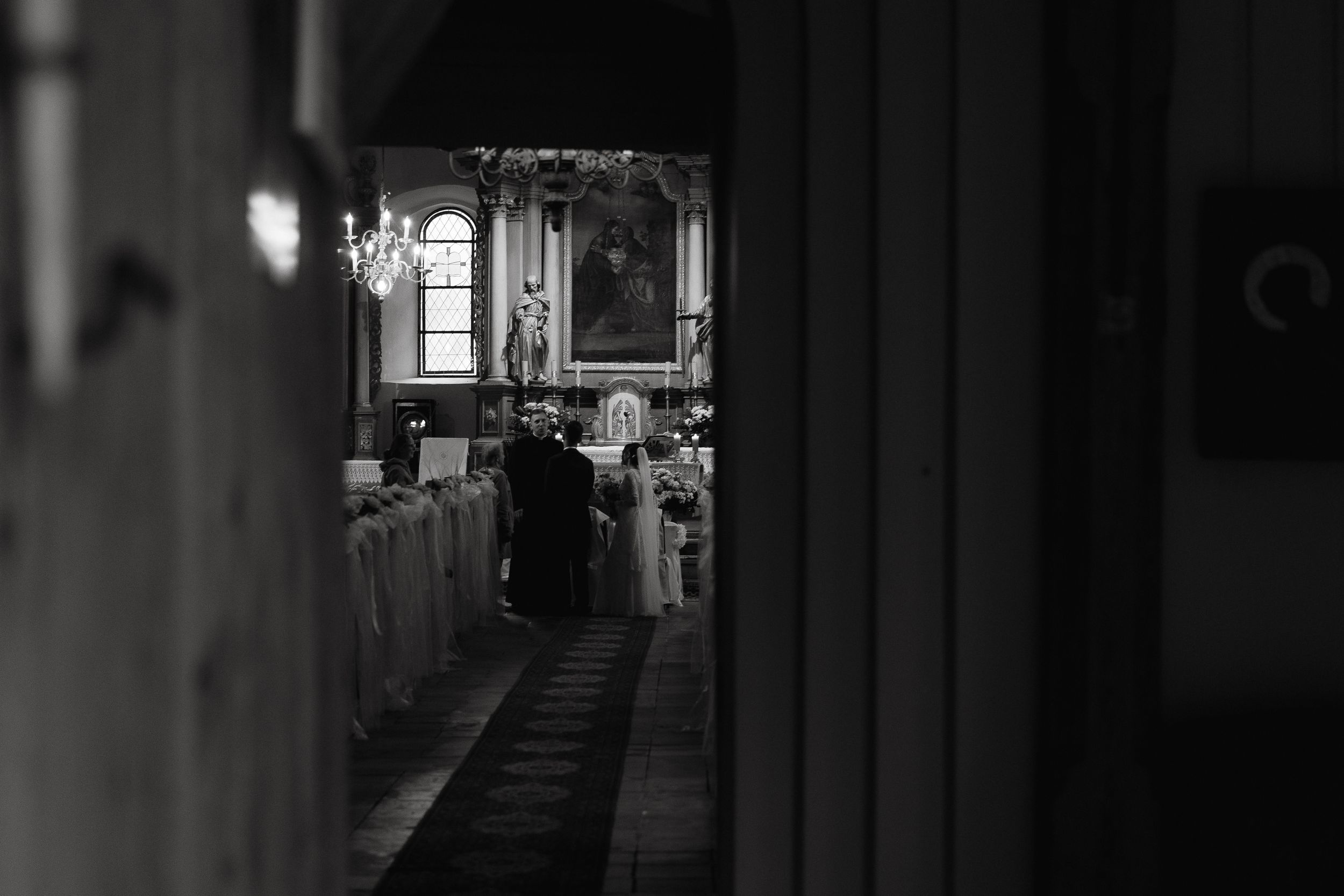 A black and white photograph showing figures walking in a church aisle between white pews.