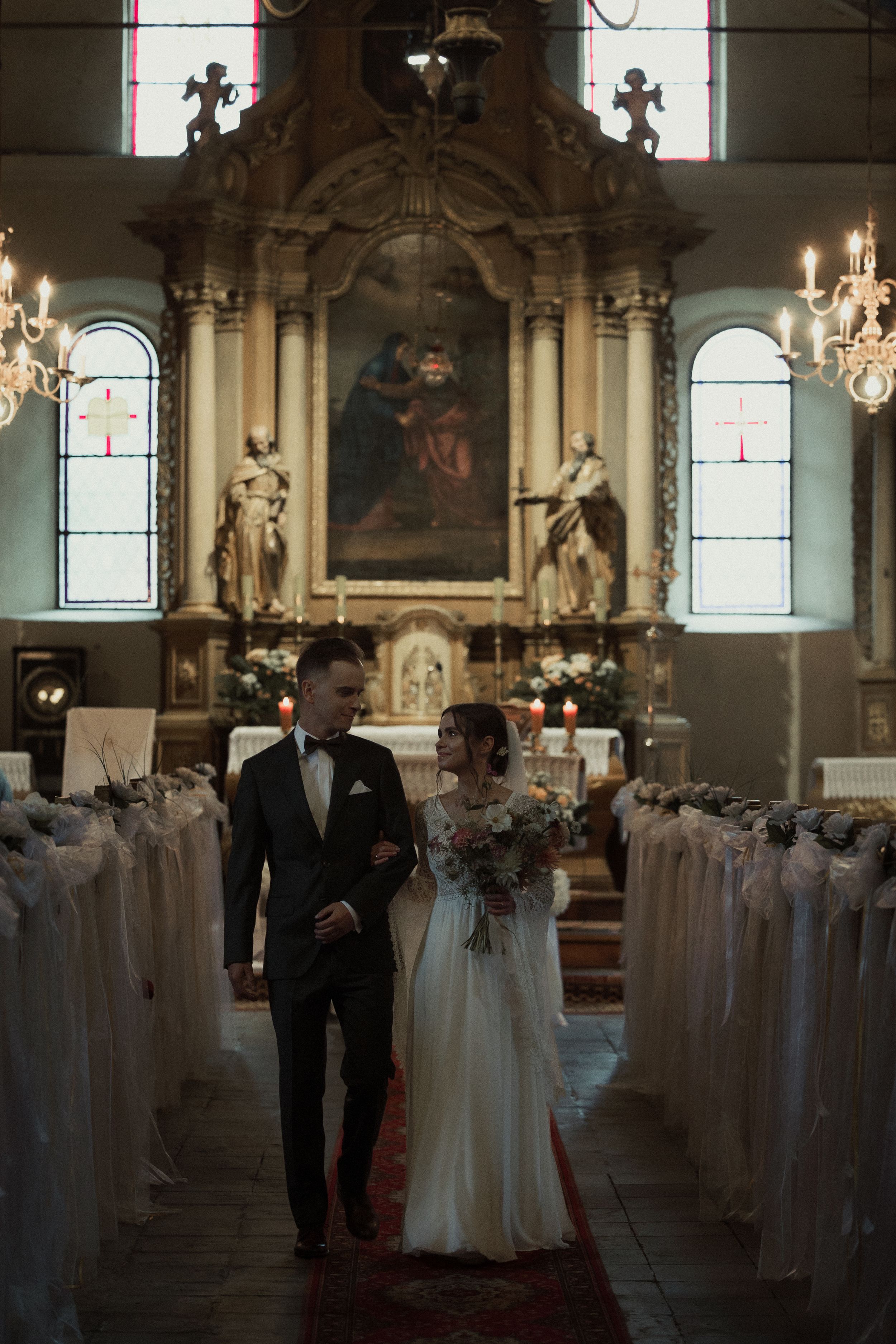 A wedding ceremony inside a grand church with rows of white-covered chairs and elegant chandeliers illuminating the space.