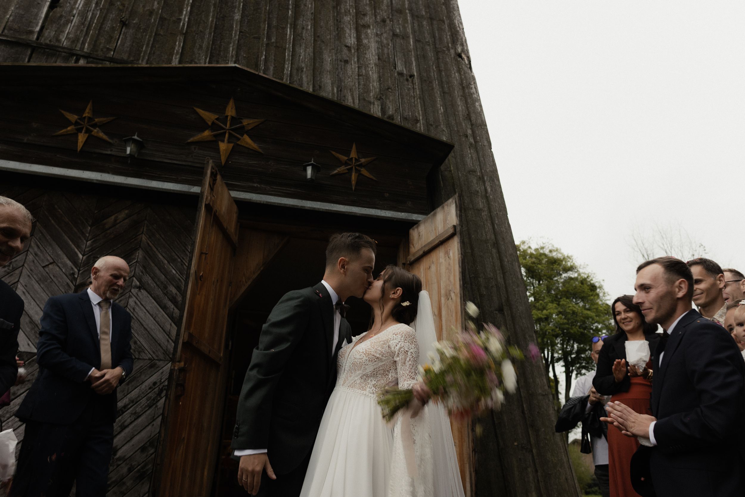 Wedding party with candid emotional reactions during ceremony under rustic wooden beams in church setting.