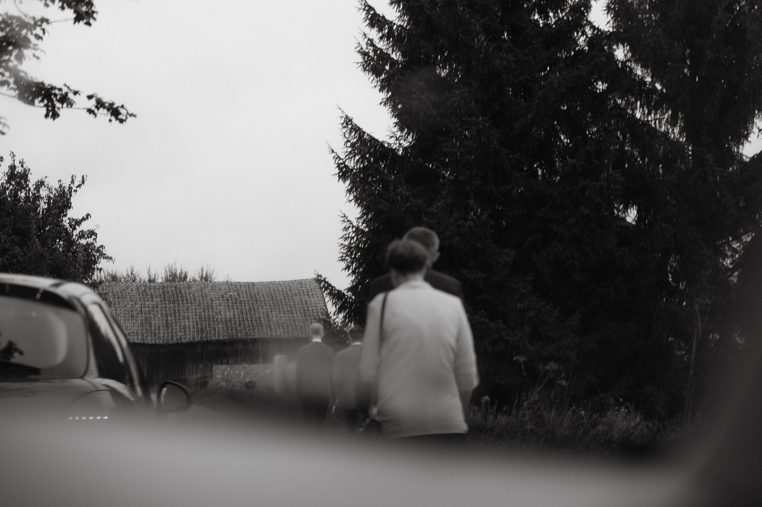 A black and white photo shot through a car window of someone in a light colored jacket sitting near evergreen trees.