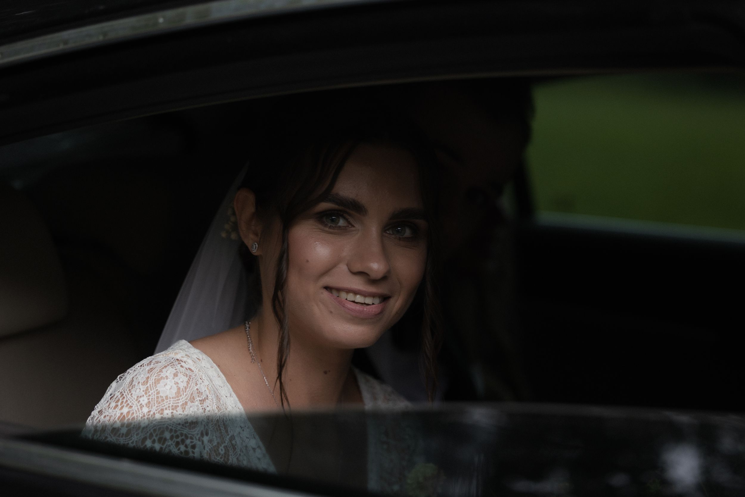 A bride smiles warmly while sitting in a car on her wedding day, wearing a white lace dress and soft veil.