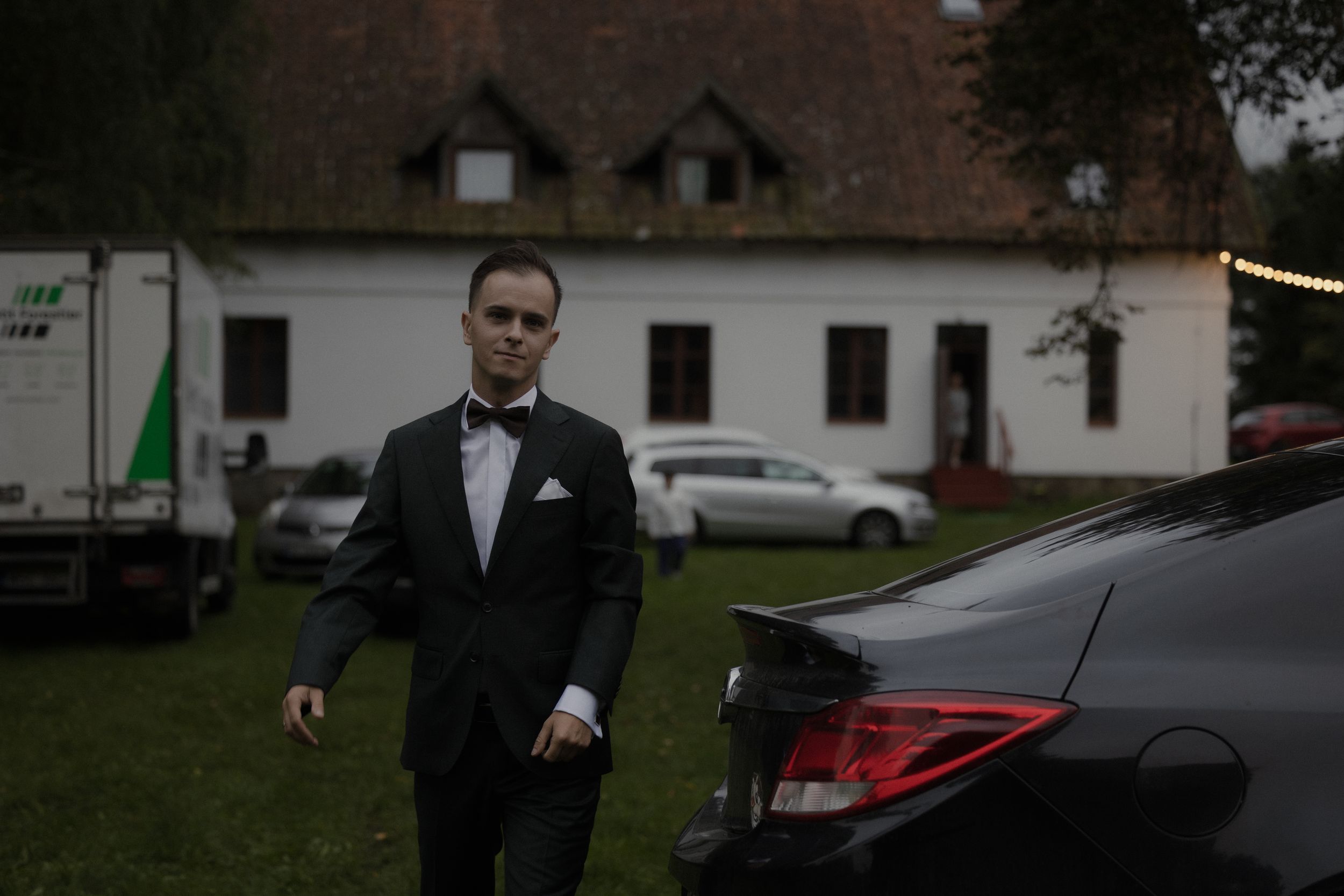 A person in a suit walks through a parking lot with buildings and trees in the background.