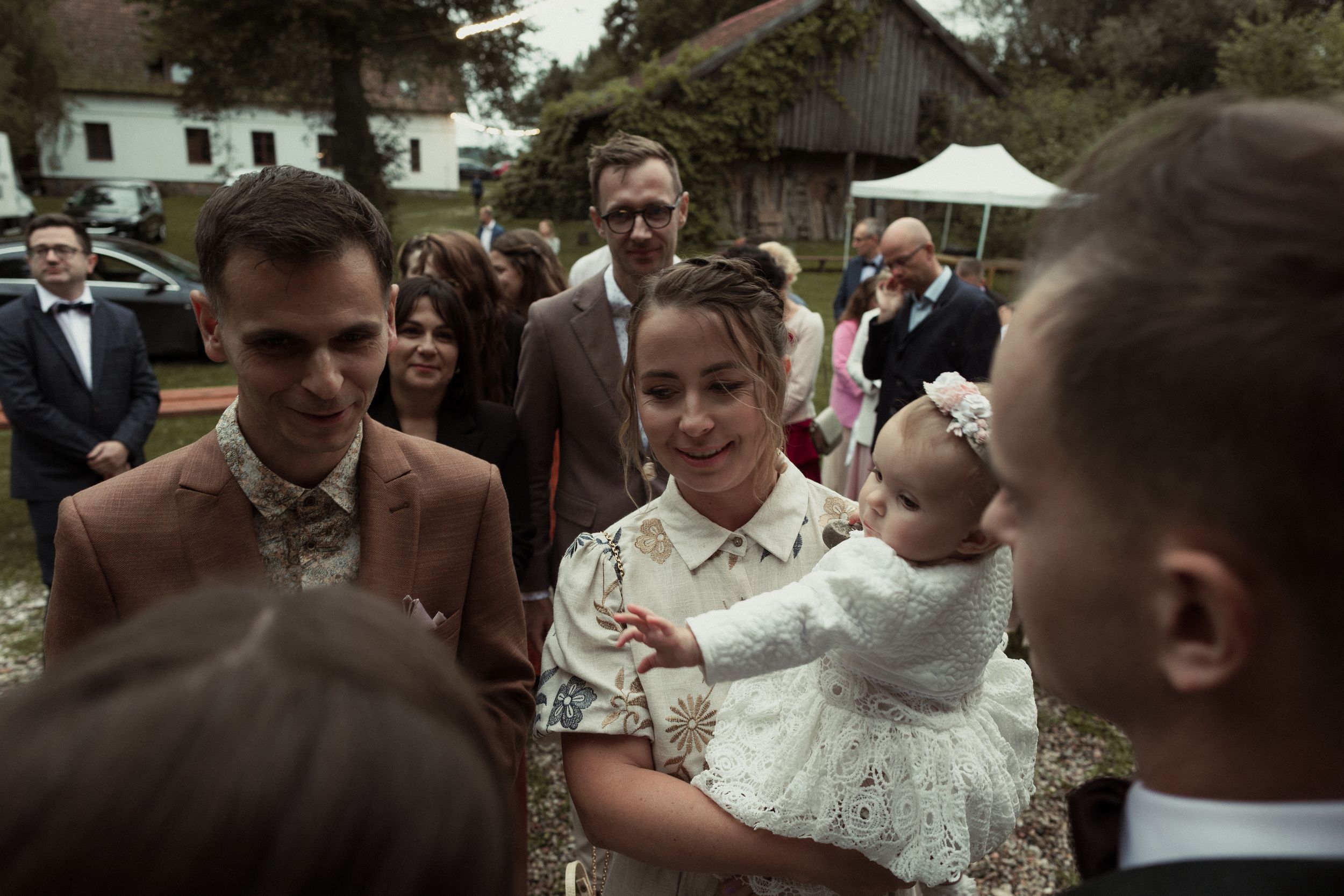 A baby in a white christening gown is held at an outdoor gathering as people in formal attire stand nearby.