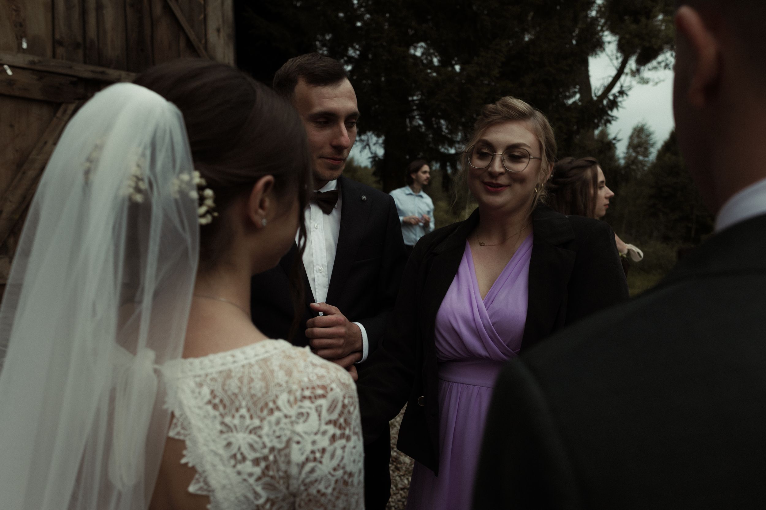 A wedding gathering outdoors shows people in formal attire including a bride in a lace dress with veil and guest in purple.