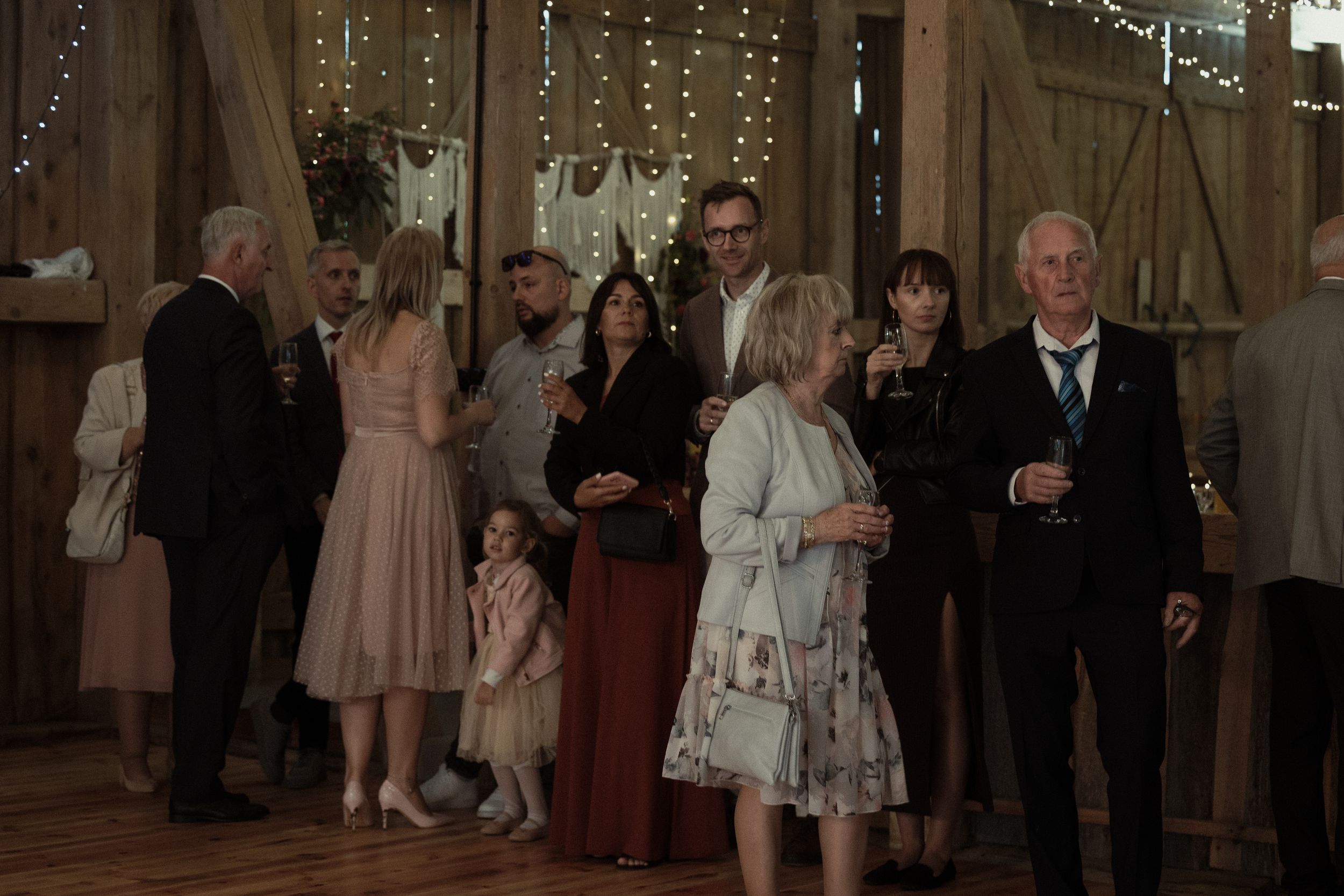 Wedding guests mingle and dance in a rustic barn reception venue decorated with twinkling string lights.