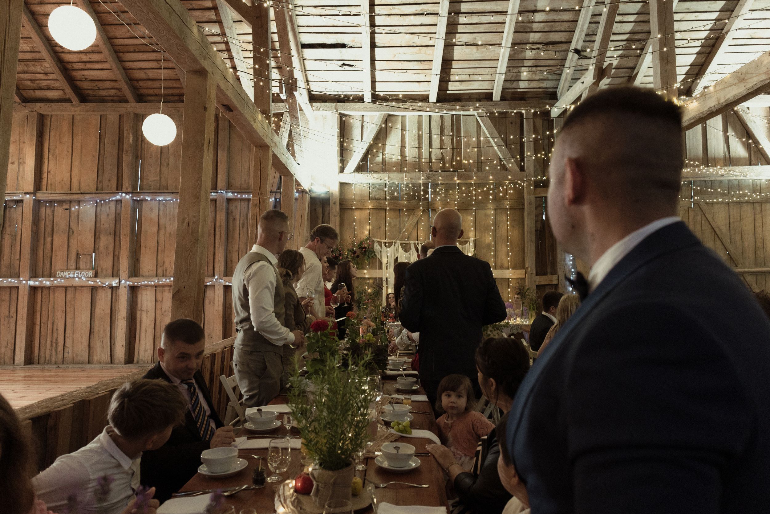 A rustic indoor barn wedding reception with wooden beams, string lights, and elegant table settings.