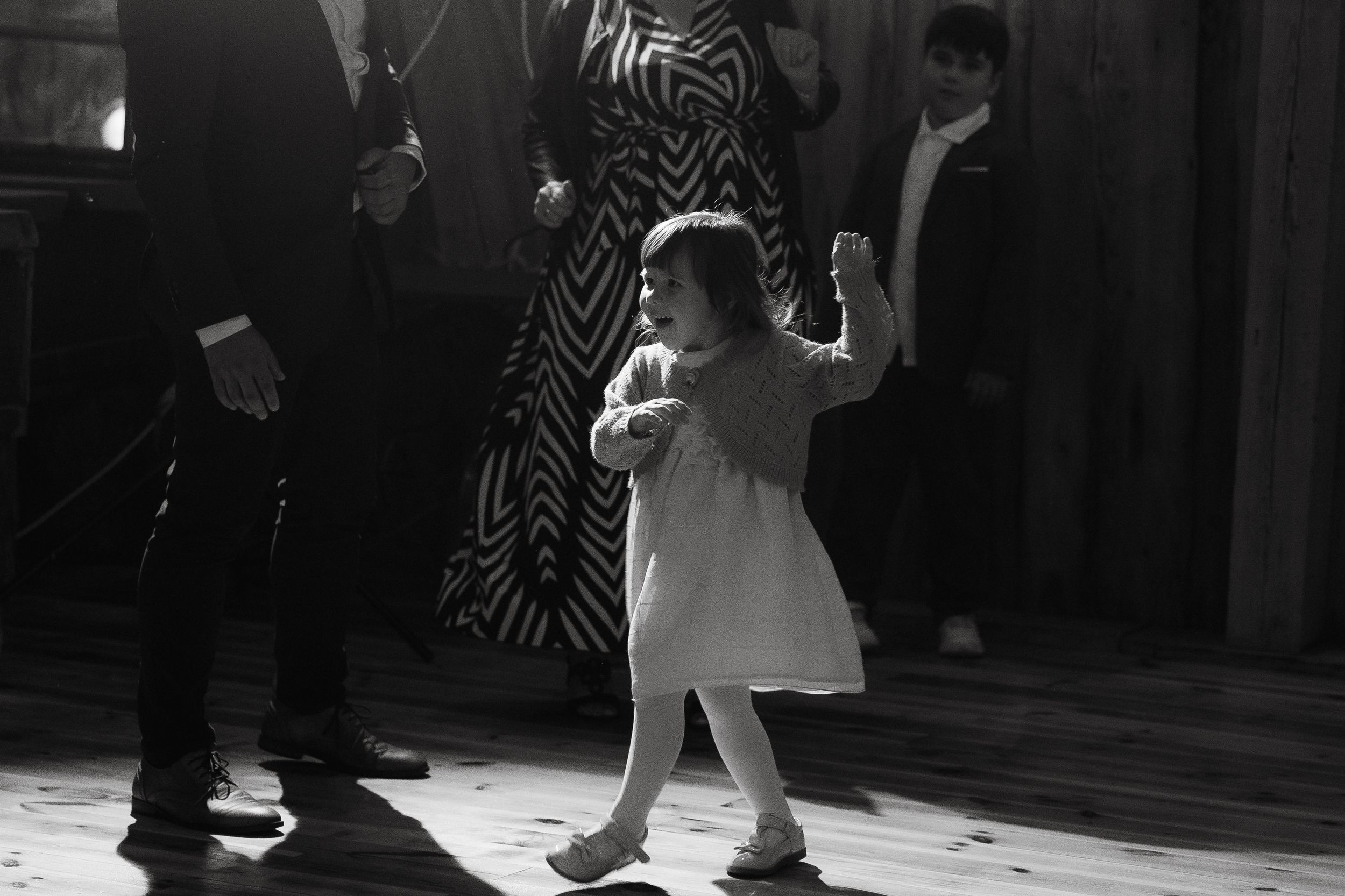 A small child in a white dress dances in a beam of light on a dark dance floor during a celebratory event.