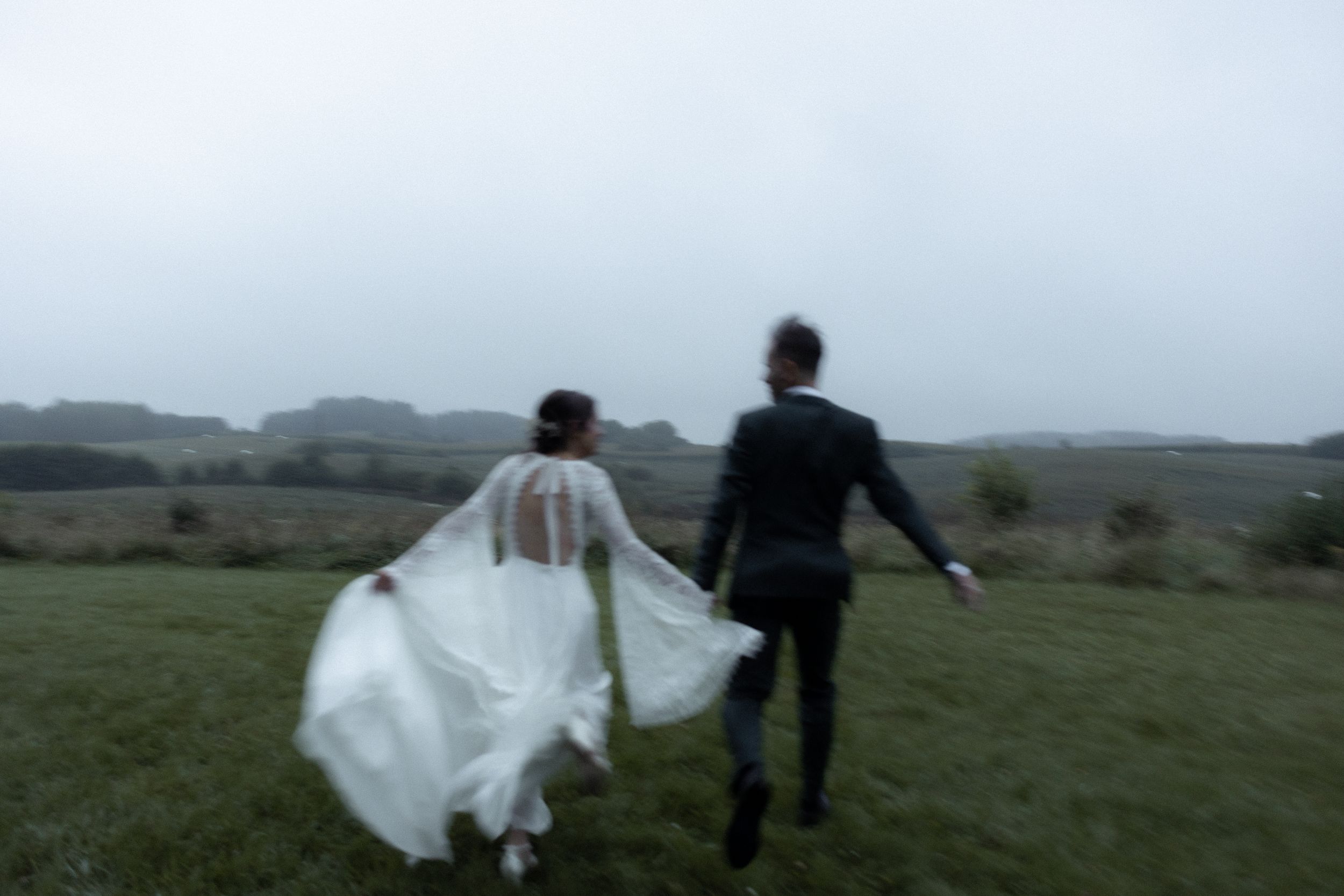 Wedding couple walks through misty green countryside meadow holding hands in silhouette at dusk.