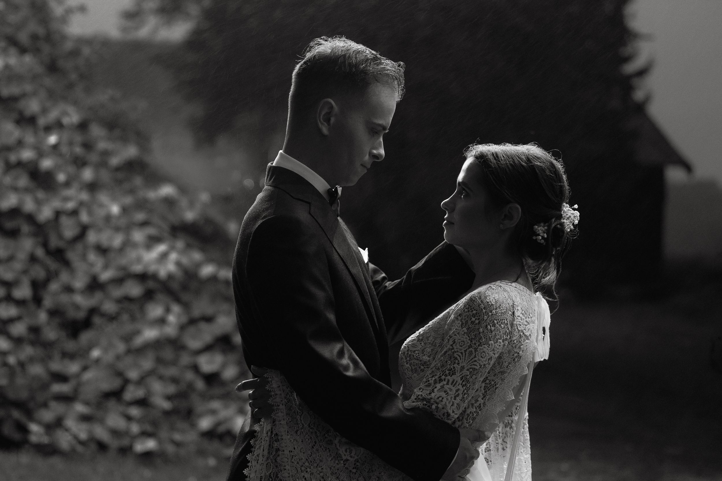 A romantic black and white wedding photo of a couple embracing in front of a garden setting.