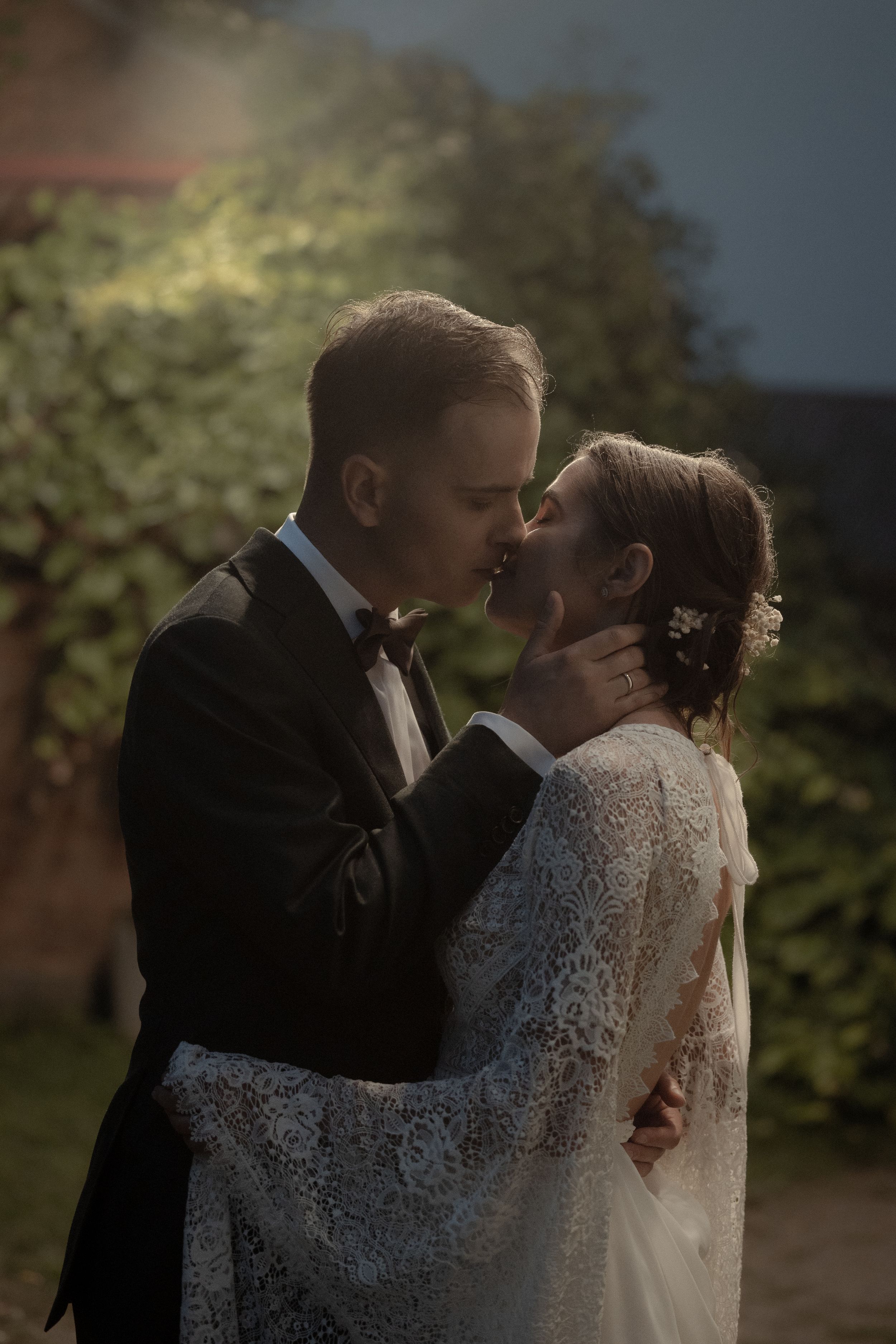 A tender wedding moment captured as a couple in formal attire share an intimate kiss in a garden.