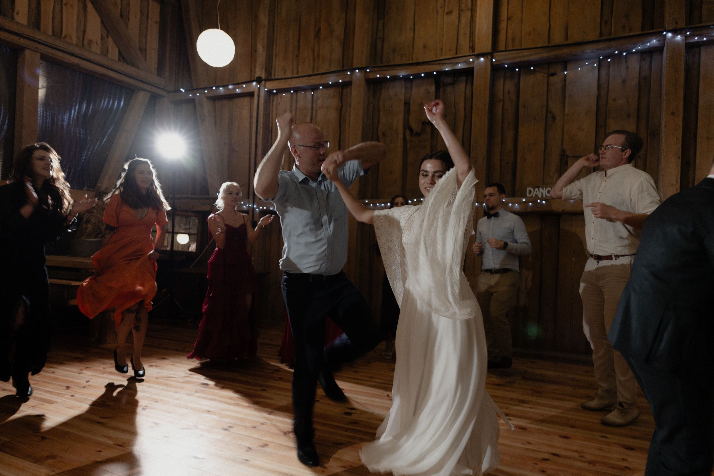 Wedding guests and a bride dance joyfully under string lights in a rustic wooden barn during the reception celebration.