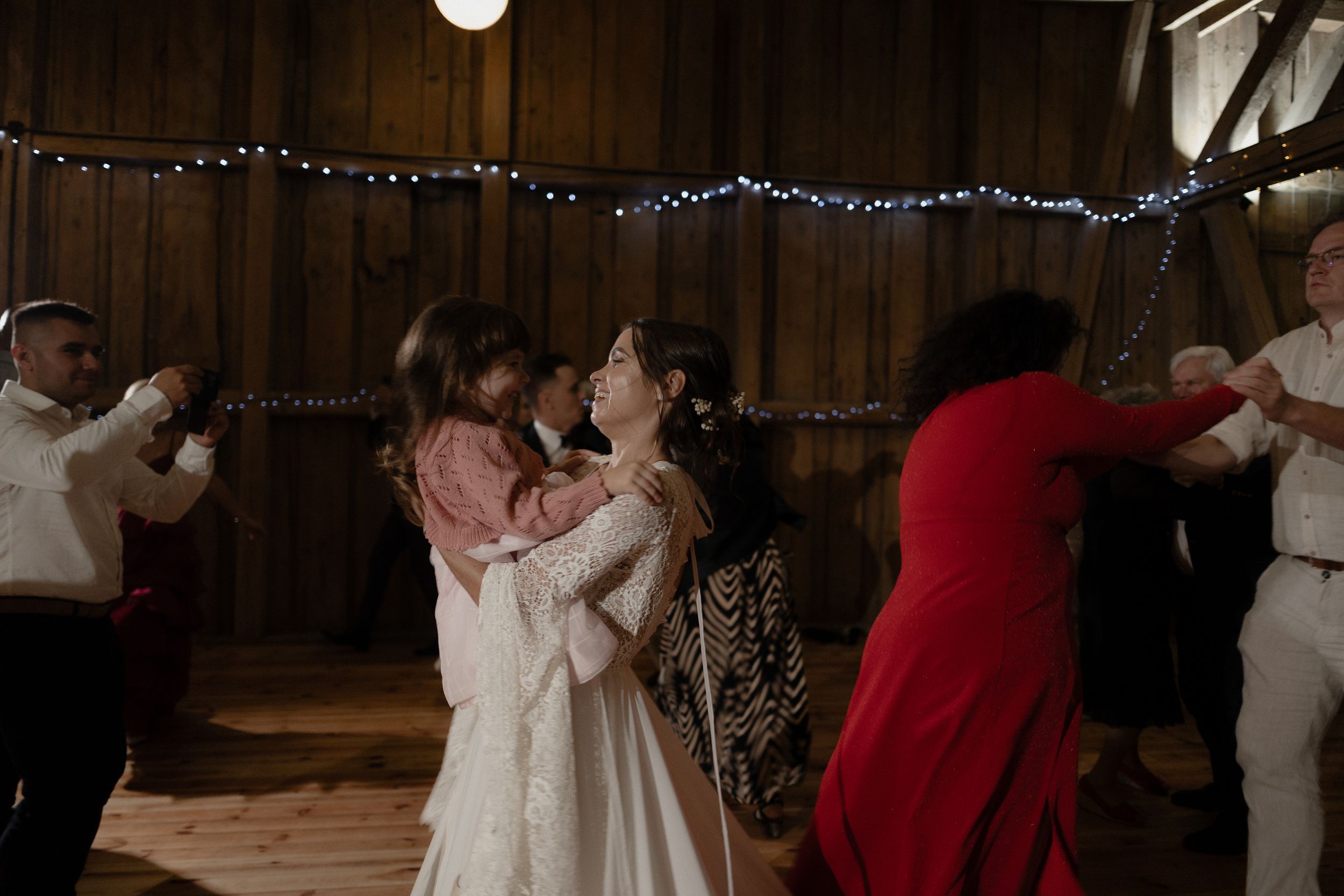 People dance and celebrate inside a rustic barn venue decorated with string lights during a wedding celebration.